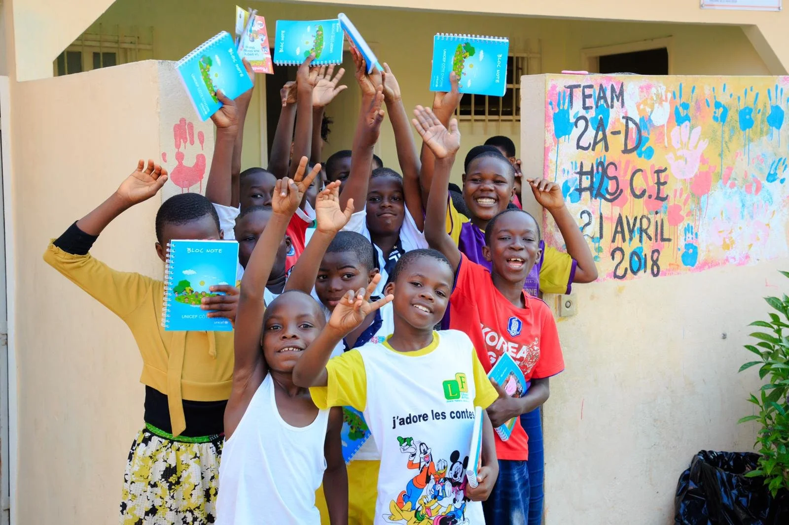 Group of children smiling and holding notebooks outdoors in front of a colorful painted wall with text, celebrating an event on April 2, 2018.