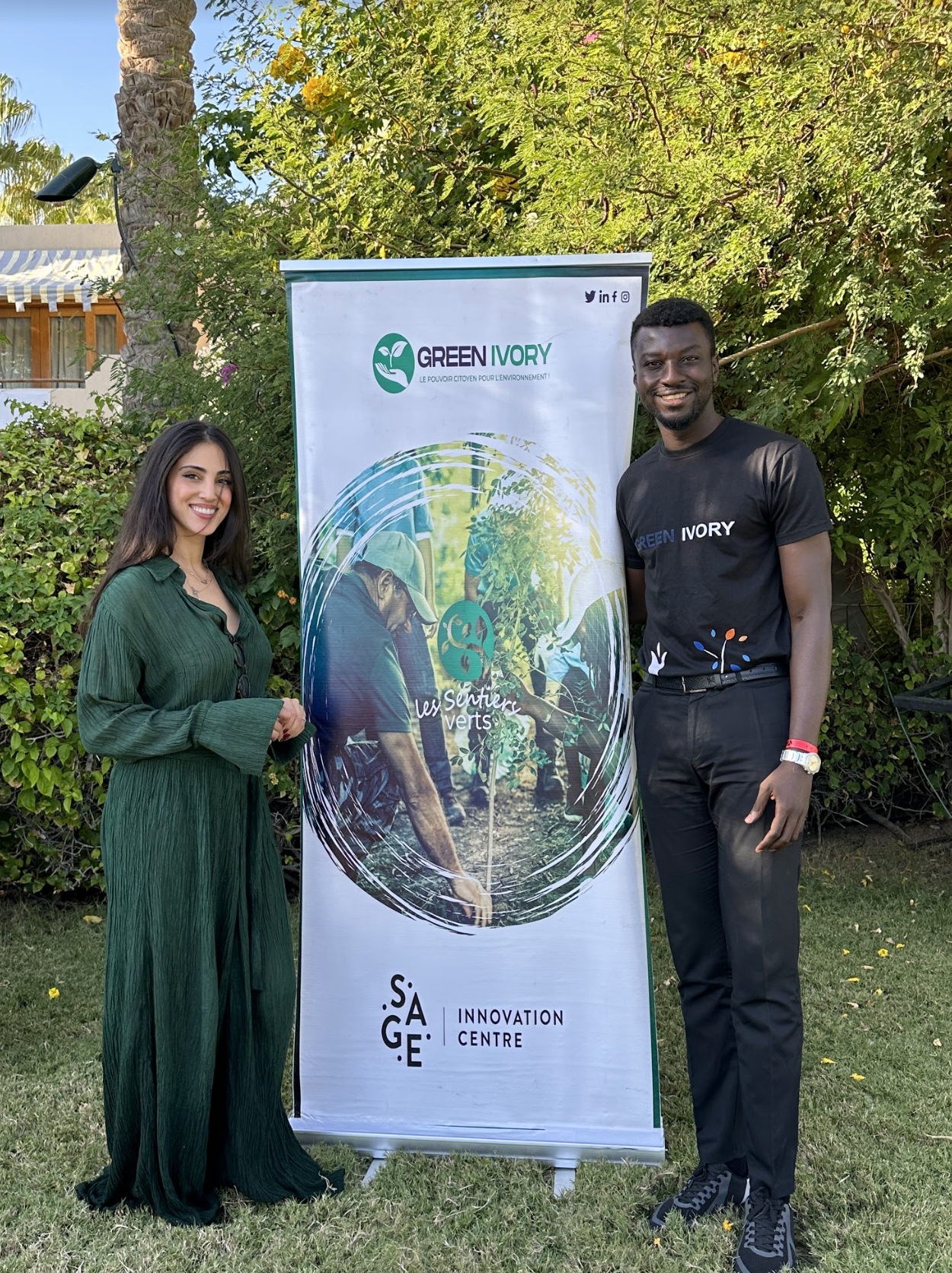 Two people, a woman and a man, standing outdoors in front of a banner for Green Ivory and Les Sentiers Verts, smiling at the camera. The woman is wearing a long green dress, and the man is dressed in a black T-shirt with the same logo and black pants. There is greenery and a house with a white awning in the background.