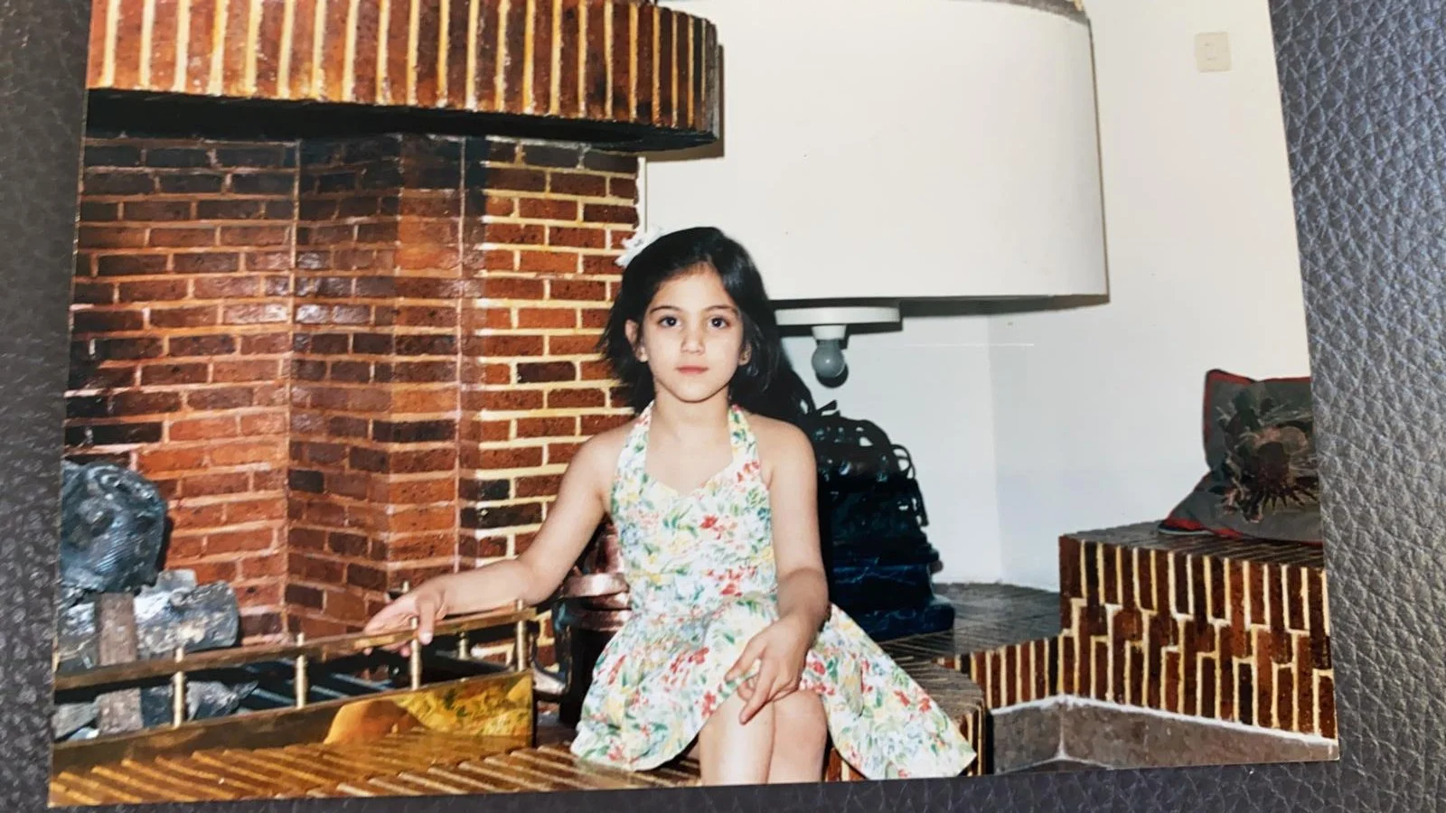 A young girl in a floral dress sitting on a brick fireplace and bench, with a pillow and black bag behind her.