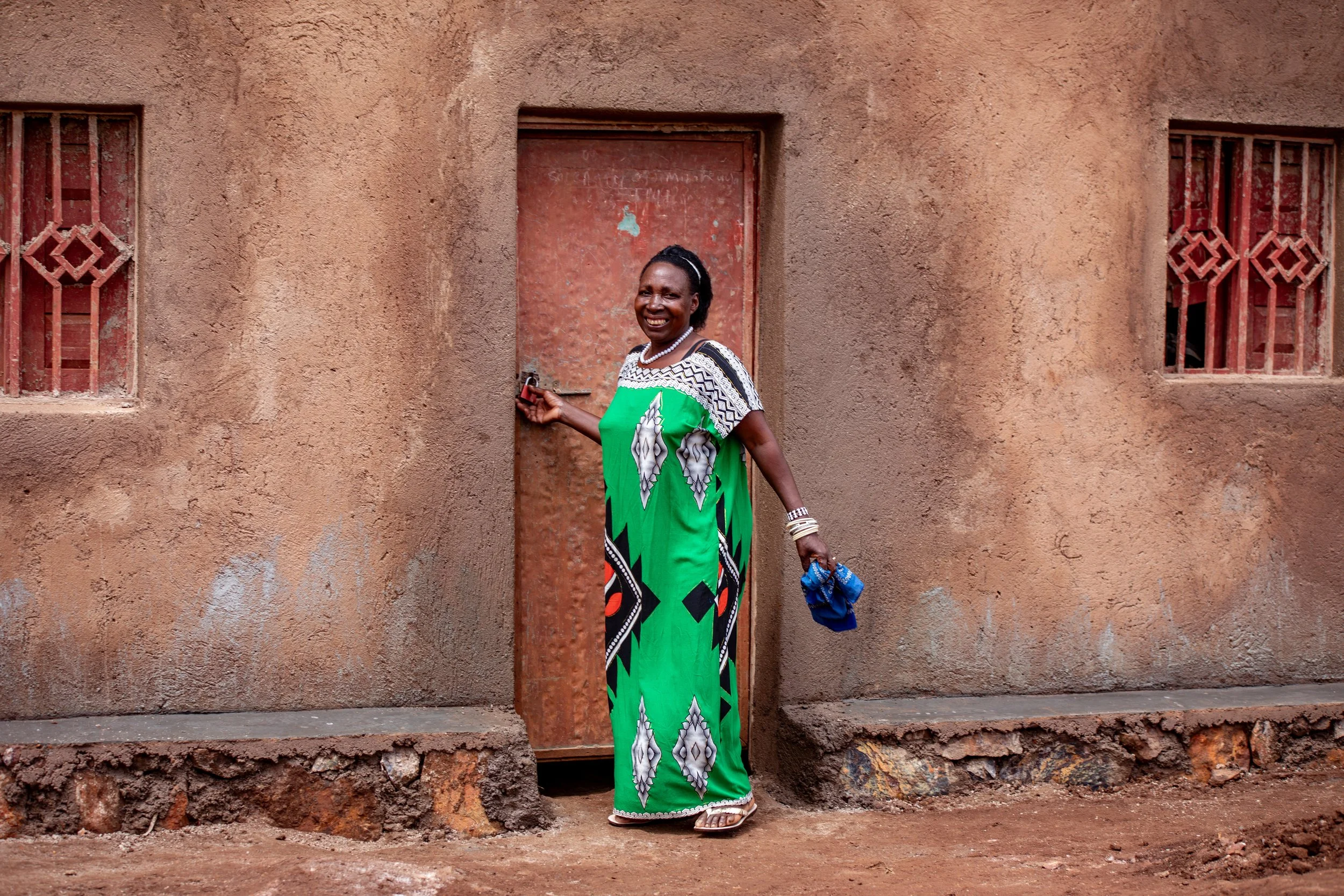 A woman smiling and standing in front of a weathered brown wall with two small barred windows and a closed red door. She is holding a key in her right hand and has a blue cloth in her left hand. The woman is wearing a colorful dress with green, black, white, and orange patterns and multiple bracelets, earrings, and a necklace.