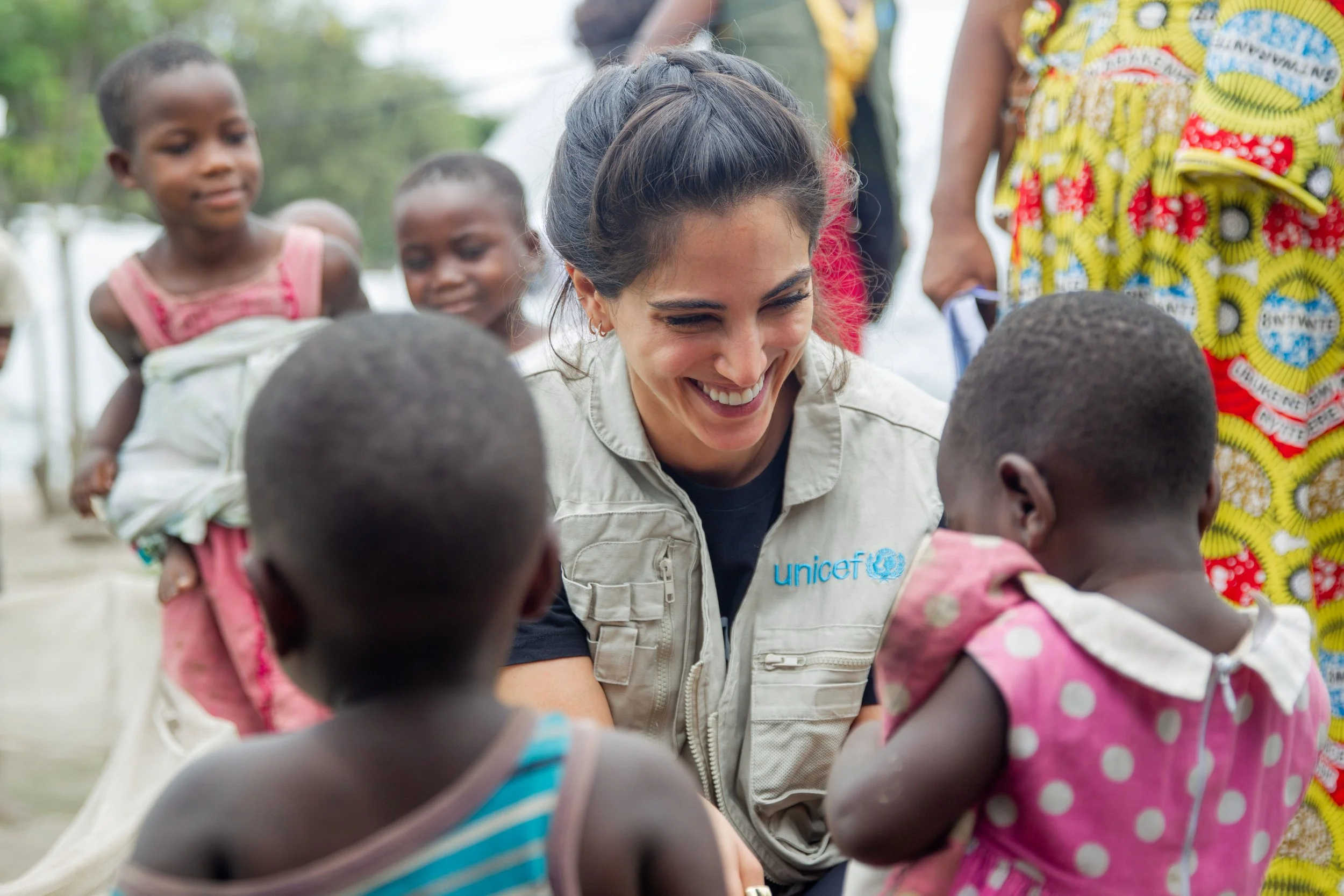 A woman wearing a UNICEF vest smiling and interacting with children in an outdoor setting with trees in the background.