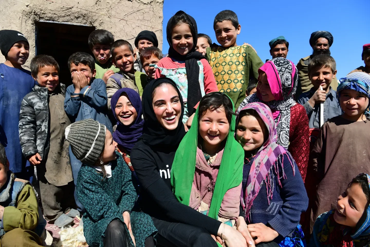 A group of smiling children and a woman sitting outdoors in front of a building under a blue sky.