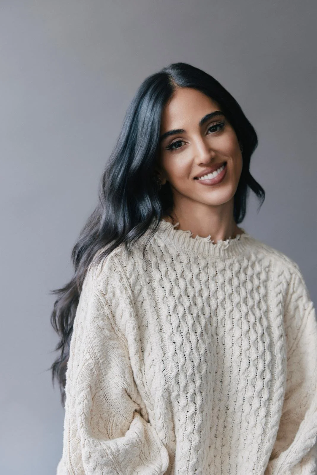A woman with long dark hair, smiling, wearing a cream-colored knit sweater, standing against a gray background.