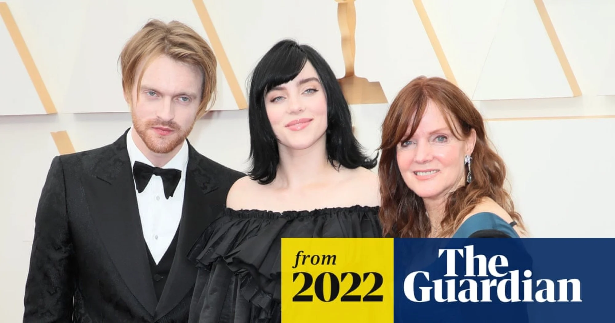 Three people in formal attire at an event, standing in front of a backdrop with geometric patterns, with a yellow and blue overlay highlighting the text 'from 2022 The Guardian'.
