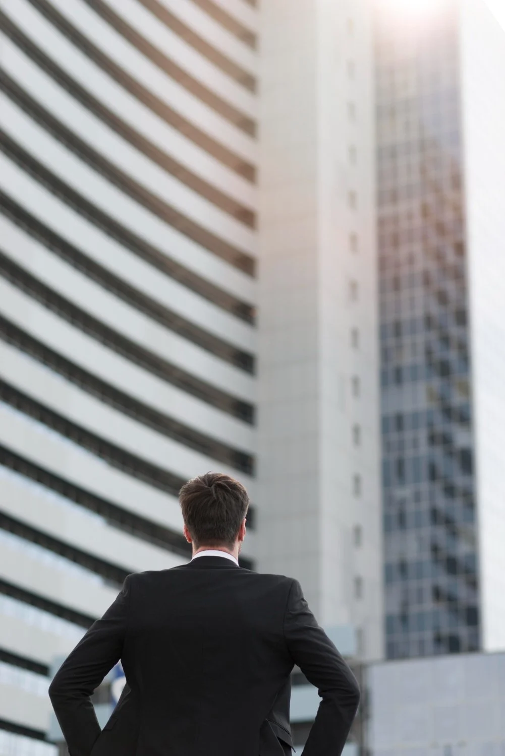 A man in a black suit standing with his back to the camera, looking up at a tall modern office building in a city.