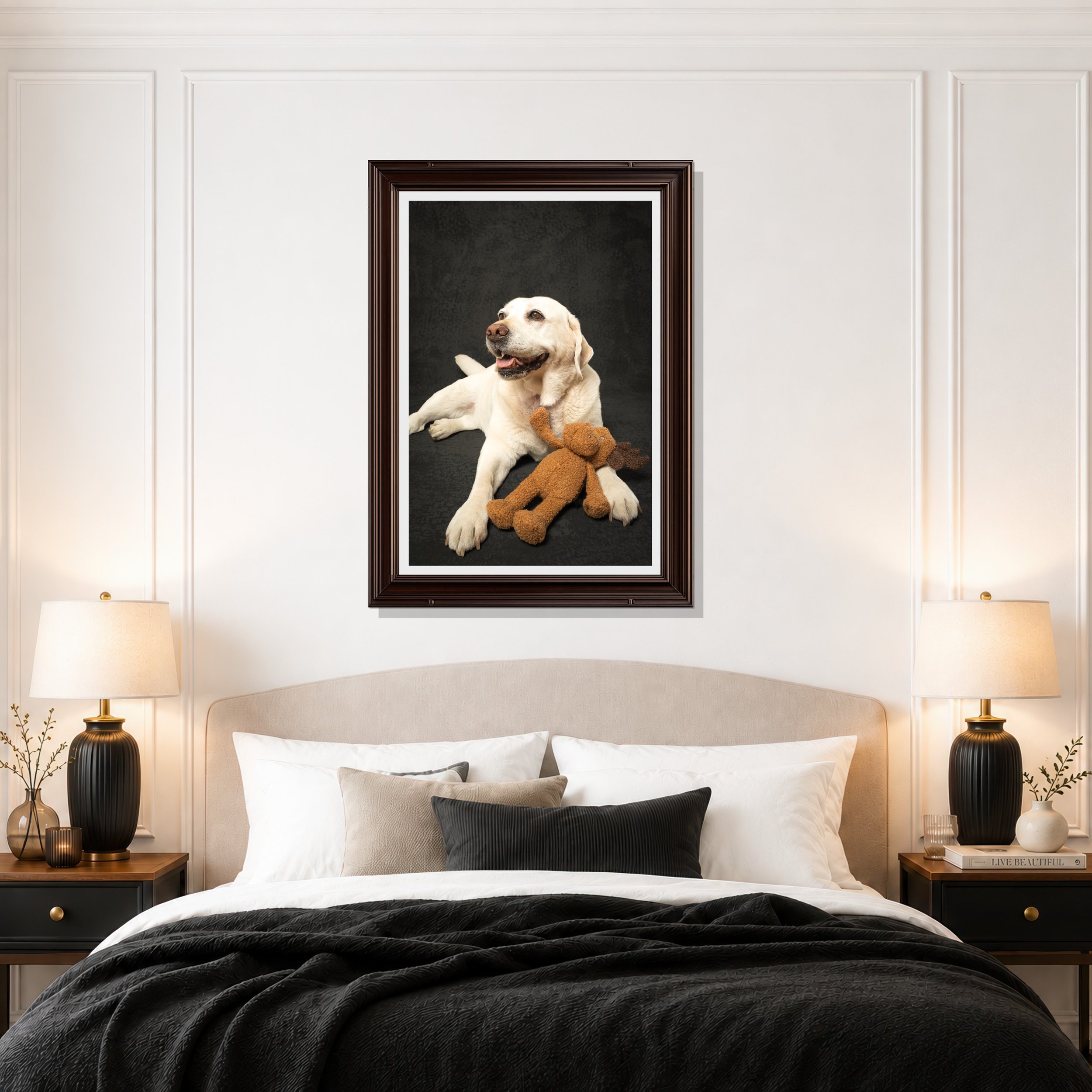 A bedroom with a large bed covered in white and black bedding, two dark nightstands with lamps, and a large portrait of a happy Labrador puppy with a teddy bear hanging out of its mouth on the wall above the bed. The room has white paneled walls and a modern decor style.