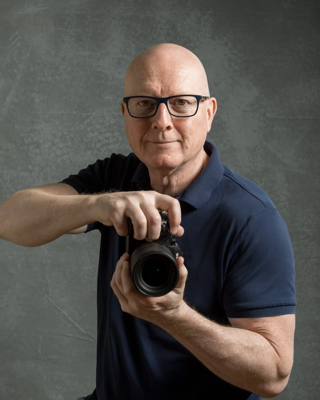 The photographer with glasses wearing a navy blue polo shirt, holding a camera in front of a plain green background.