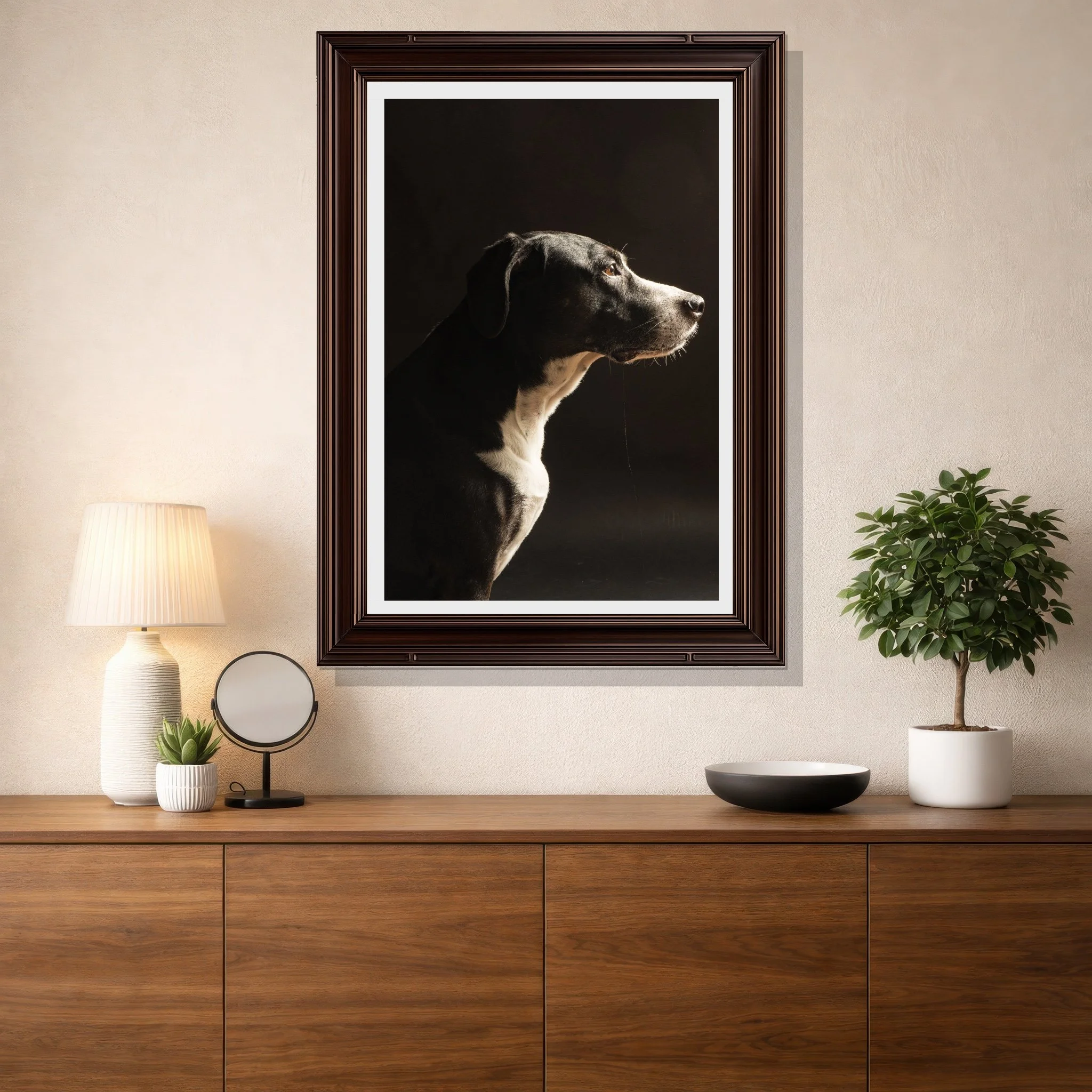 A black and white dog looking to the right, framed and hanging on a wall in a room with a wooden sideboard, a lamp, a round mirror, and a potted plant.