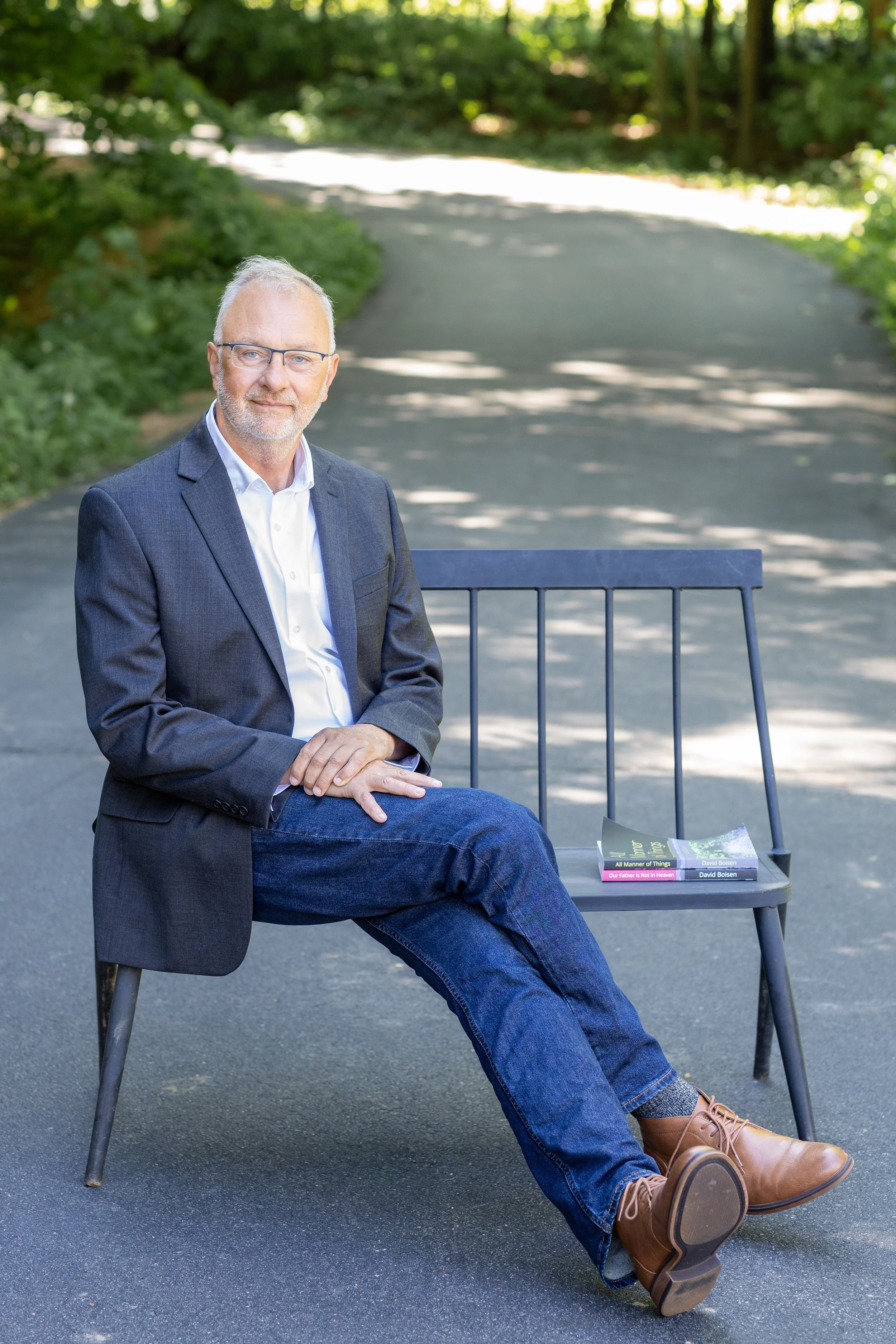 A man in a suit jacket and jeans, wearing glasses, sitting on a park bench, with two books placed on the bench next to him. The background is a shaded, wooded park area.