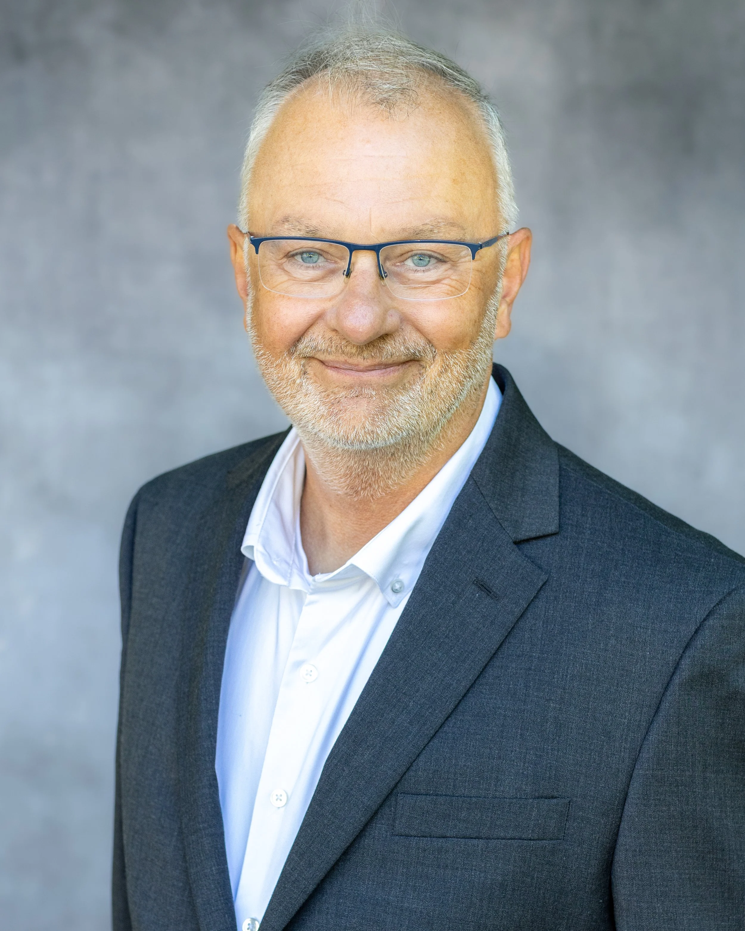 A professional headshot of David Boisen, retired Army Major and leadership speaker, with glasses, light-colored hair and beard, wearing a dark suit jacket and white dress shirt, smiling against a gray background.