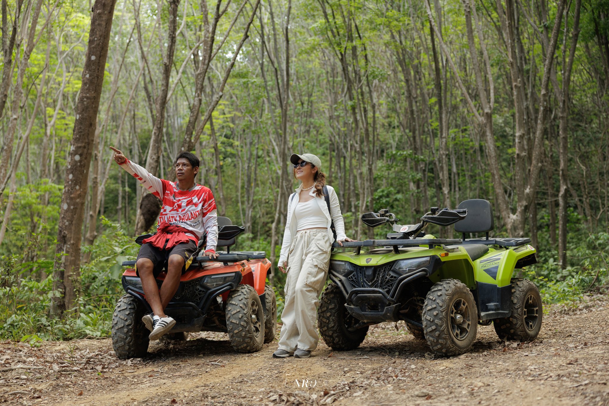 Two people, a man and a woman, in a forest, standing near and sitting on all-terrain vehicles (ATVs). The man is sitting on an orange ATV, pointing into the distance, while the woman stands beside a green ATV, smiling and looking in the same directio