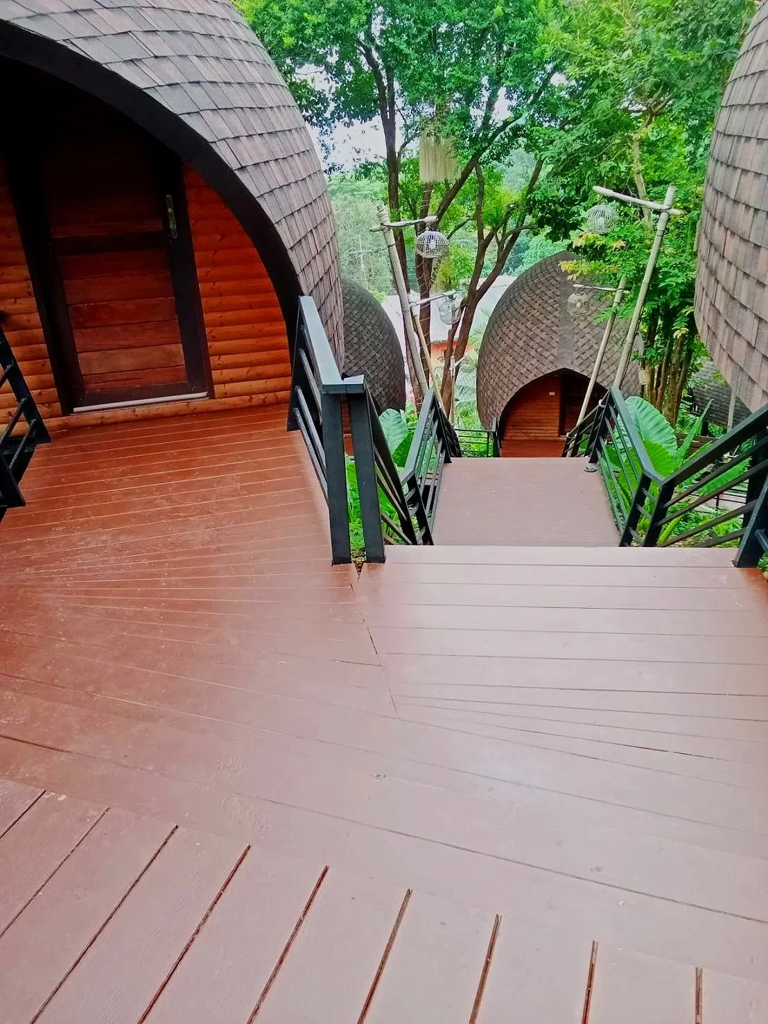 Wooden steps with black metal railings leading down to rustic, round, wooden cabins surrounded by lush green trees.