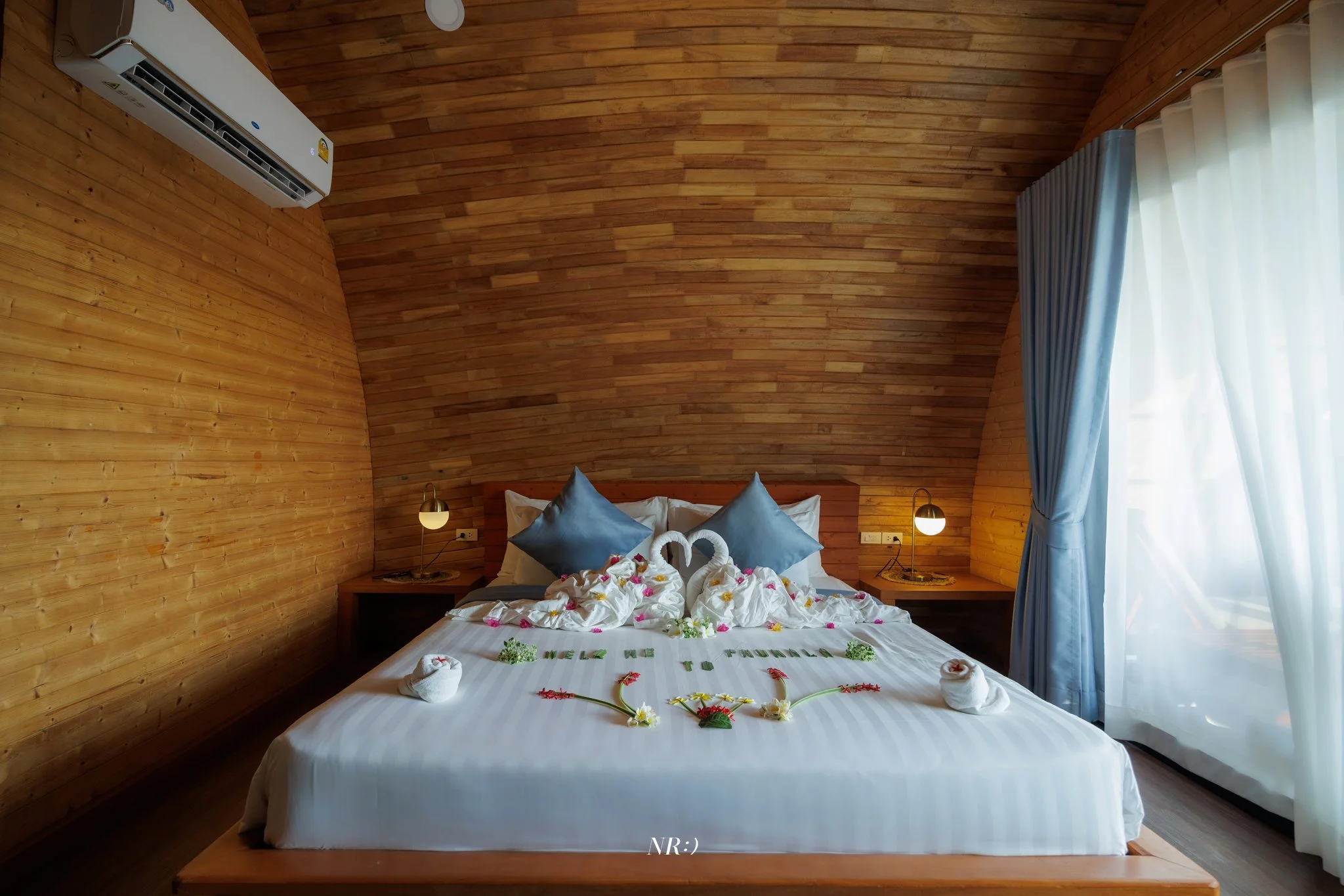 A hotel room with wooden walls and ceiling, a bed decorated with towel swans, flower petals, and a 'Welcome' message, flanked by bedside lamps and curtains on the right.