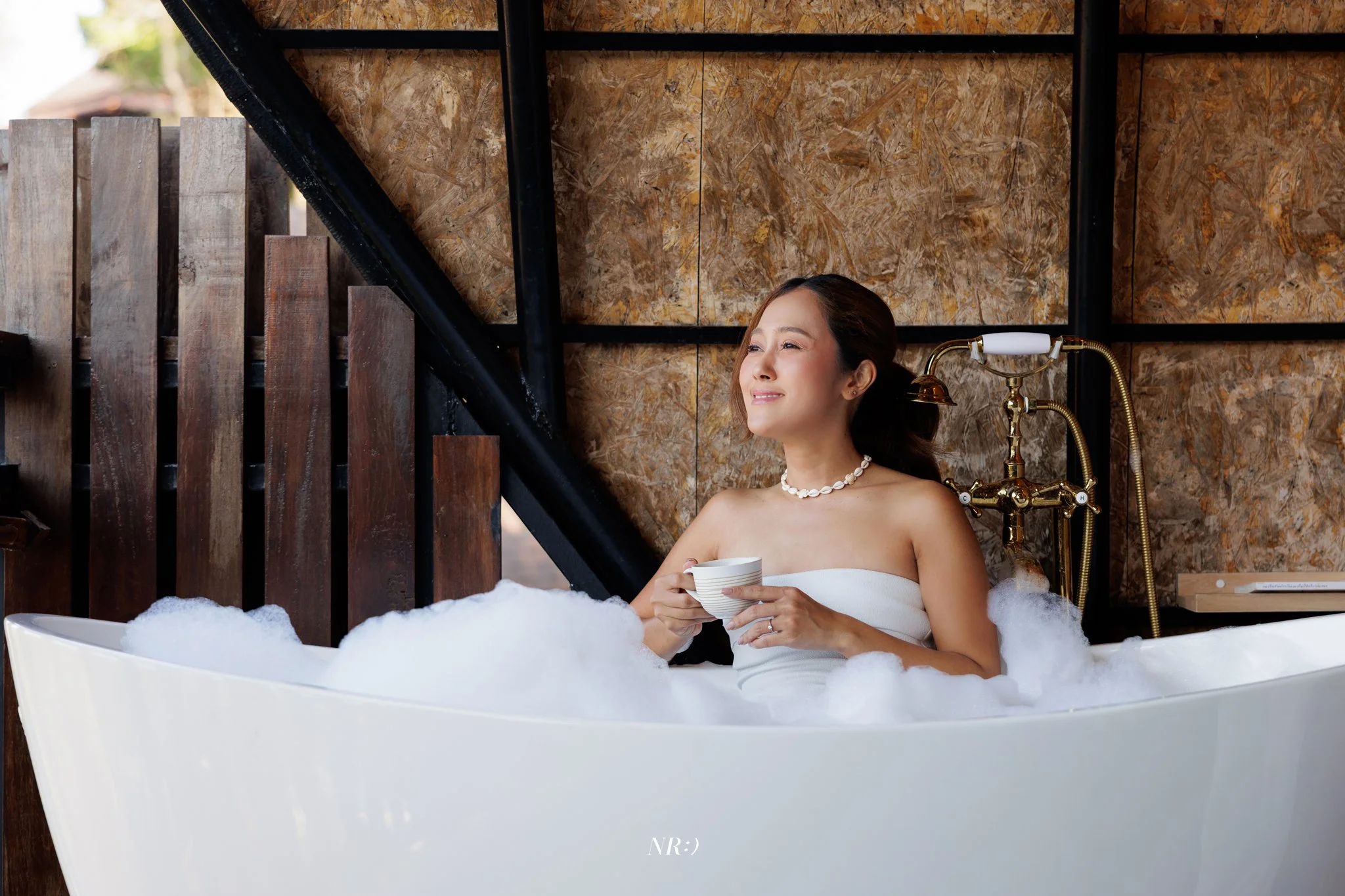 A woman relaxing in a bathtub filled with bubbles, holding a white mug, with a wooden and stone interior background, and a vintage gold faucet.