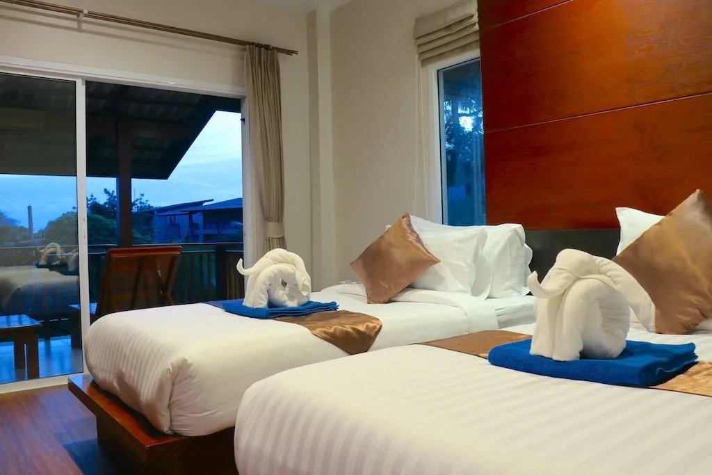 Two neatly made beds in a hotel room with towels folded into swan shapes on each bed, featuring gold and white pillows. A wooden headboard and large windows with a sliding door lead to a balcony with outdoor furniture, overlooking trees and buildings at dusk.
