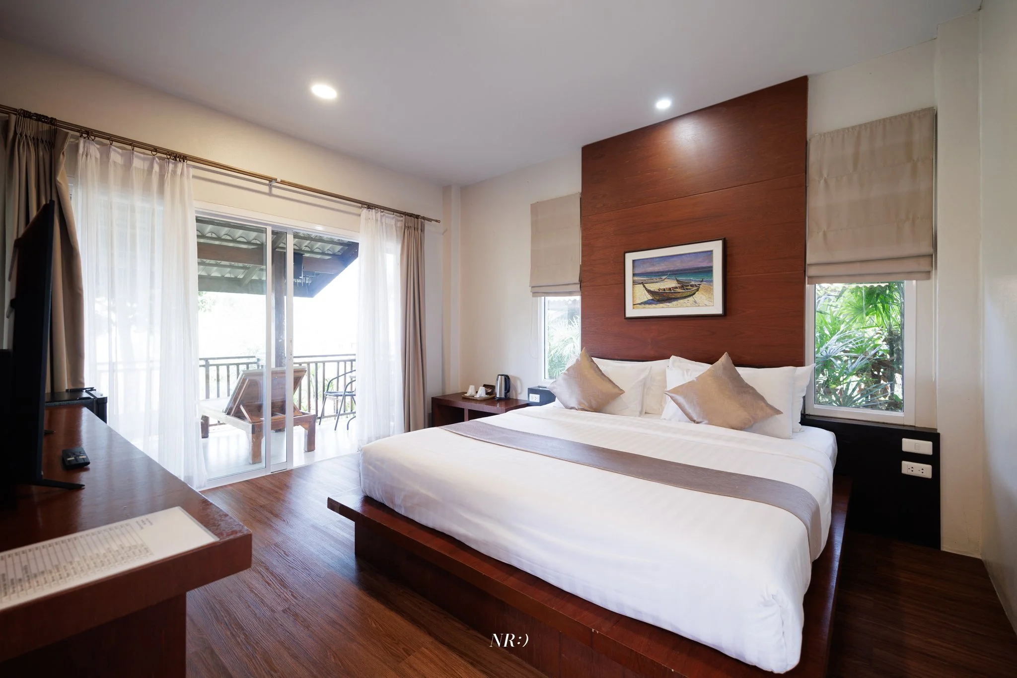 Hotel room with a bed, wooden headboard, painting of boats on the wall, large windows with beige curtains, balcony with outdoor furniture, and wooden flooring.