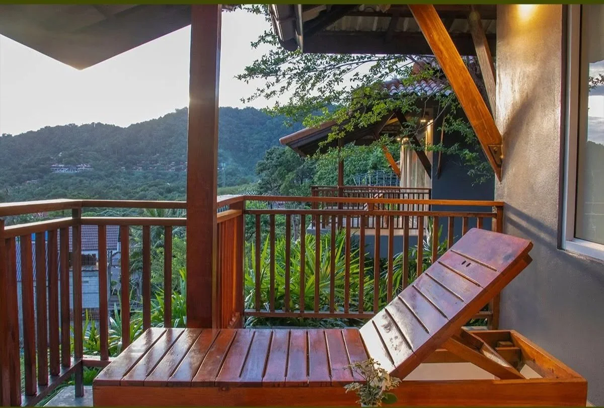 Wooden lounge chair on a balcony with a view of lush green mountains and trees in the background.