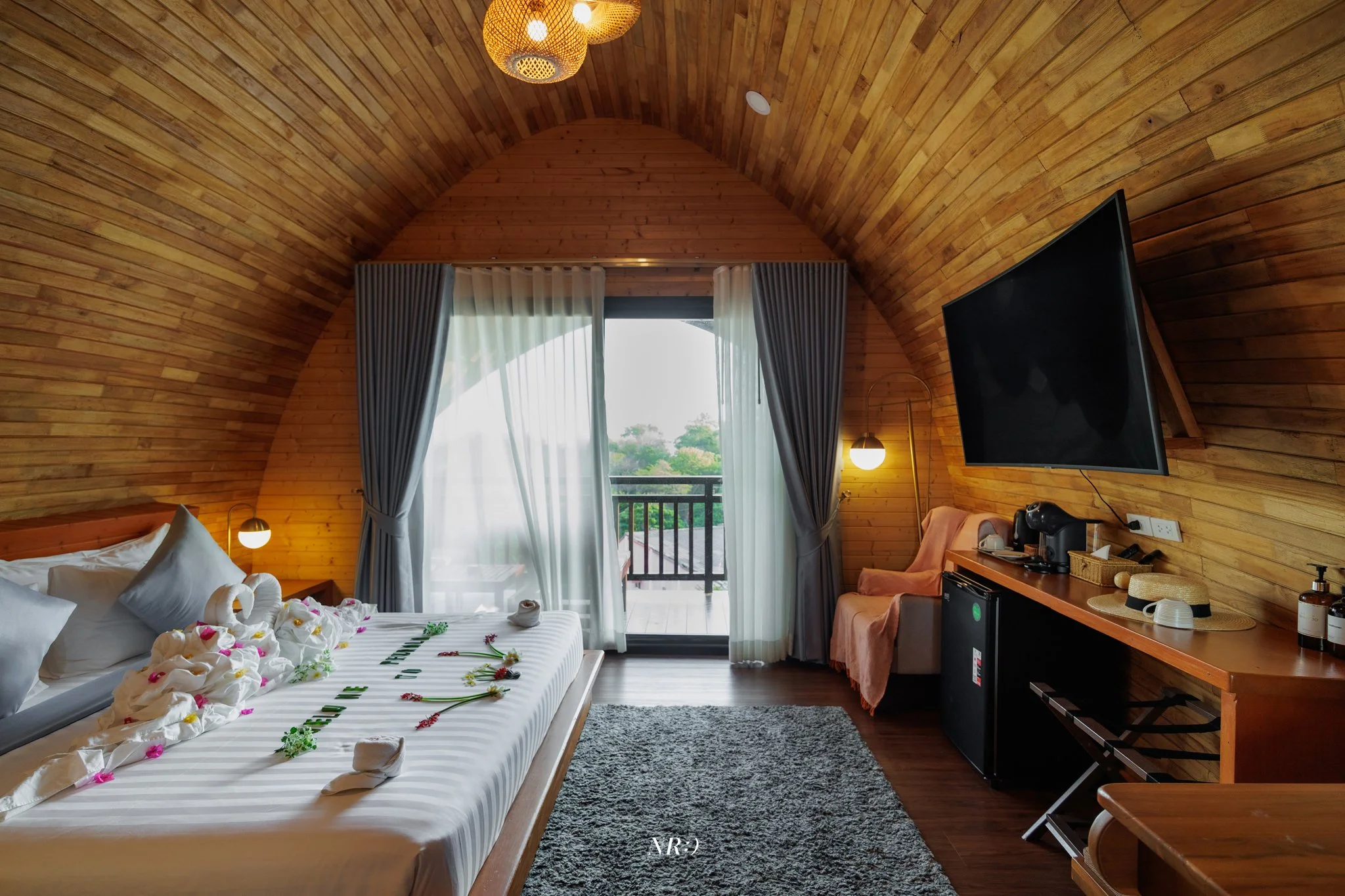 A cozy bedroom with wooden paneled walls and ceiling, a bed decorated with towel animals and flowers, a large window with white curtains, a flat-screen TV on the wall, a small balcony outside, and various furniture including a orange sofa, a wooden d
