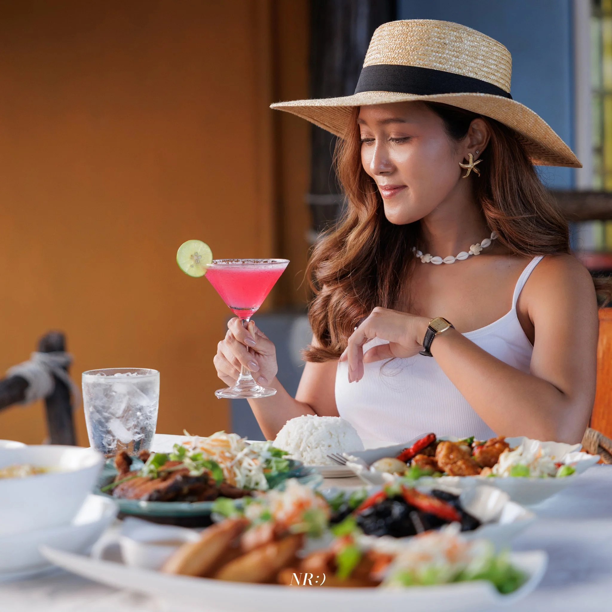 A woman wearing a large straw hat, starfish earrings, and a shell necklace, holding a pink cocktail with a cucumber slice garnish, seated at a table with a spread of food including rice, vegetables, and meat.