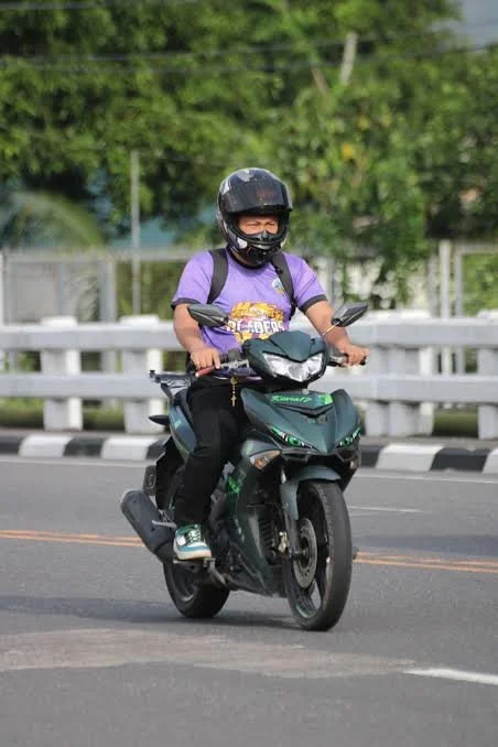 A person riding a black and green electric scooter on a road, wearing a black helmet, purple t-shirt, black pants, and sneakers.