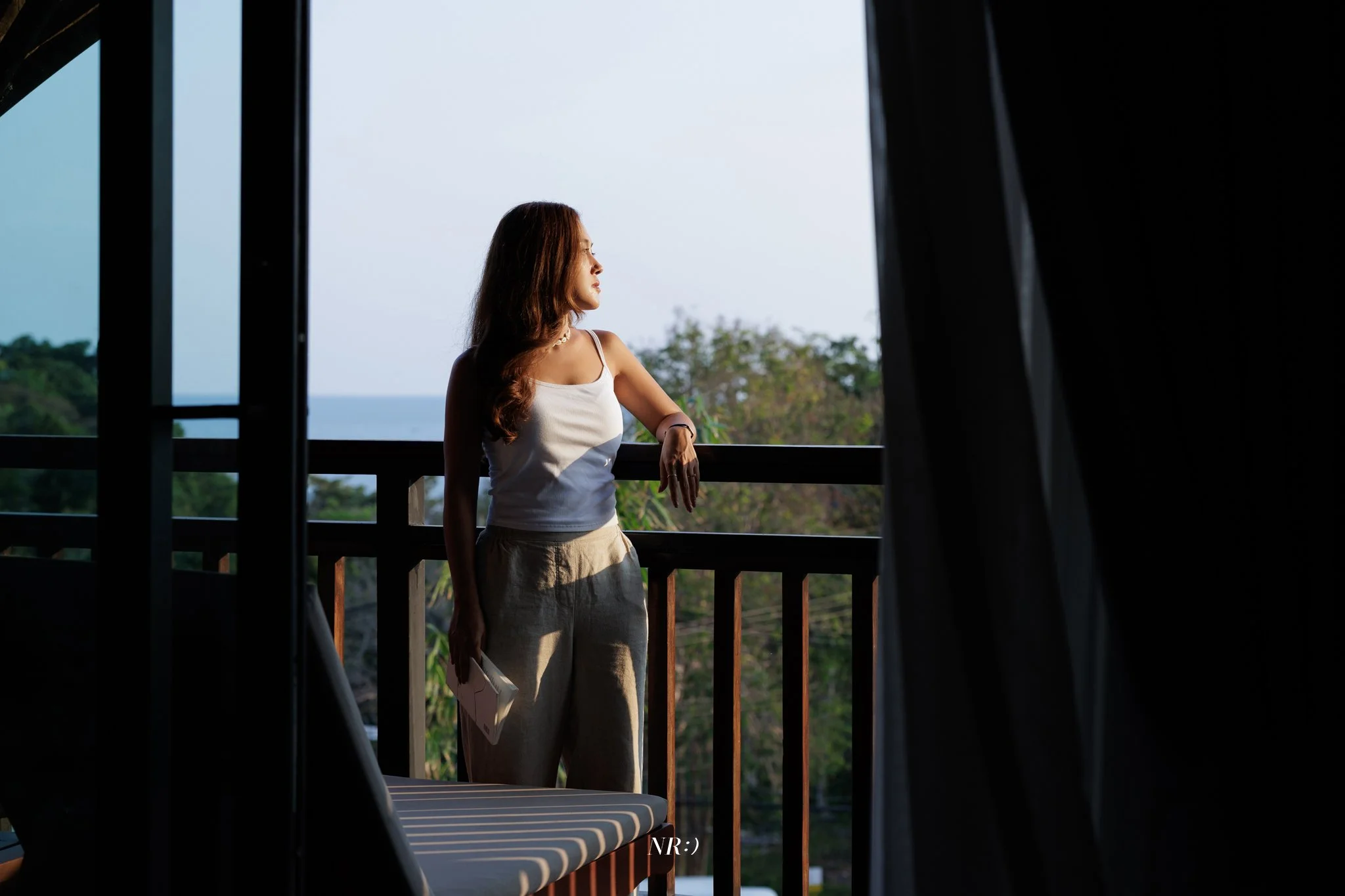 A balcony, gazing out at the ocean and greenery in the distance during daytime. The balcony has a wooden railing and a curtain on one side.