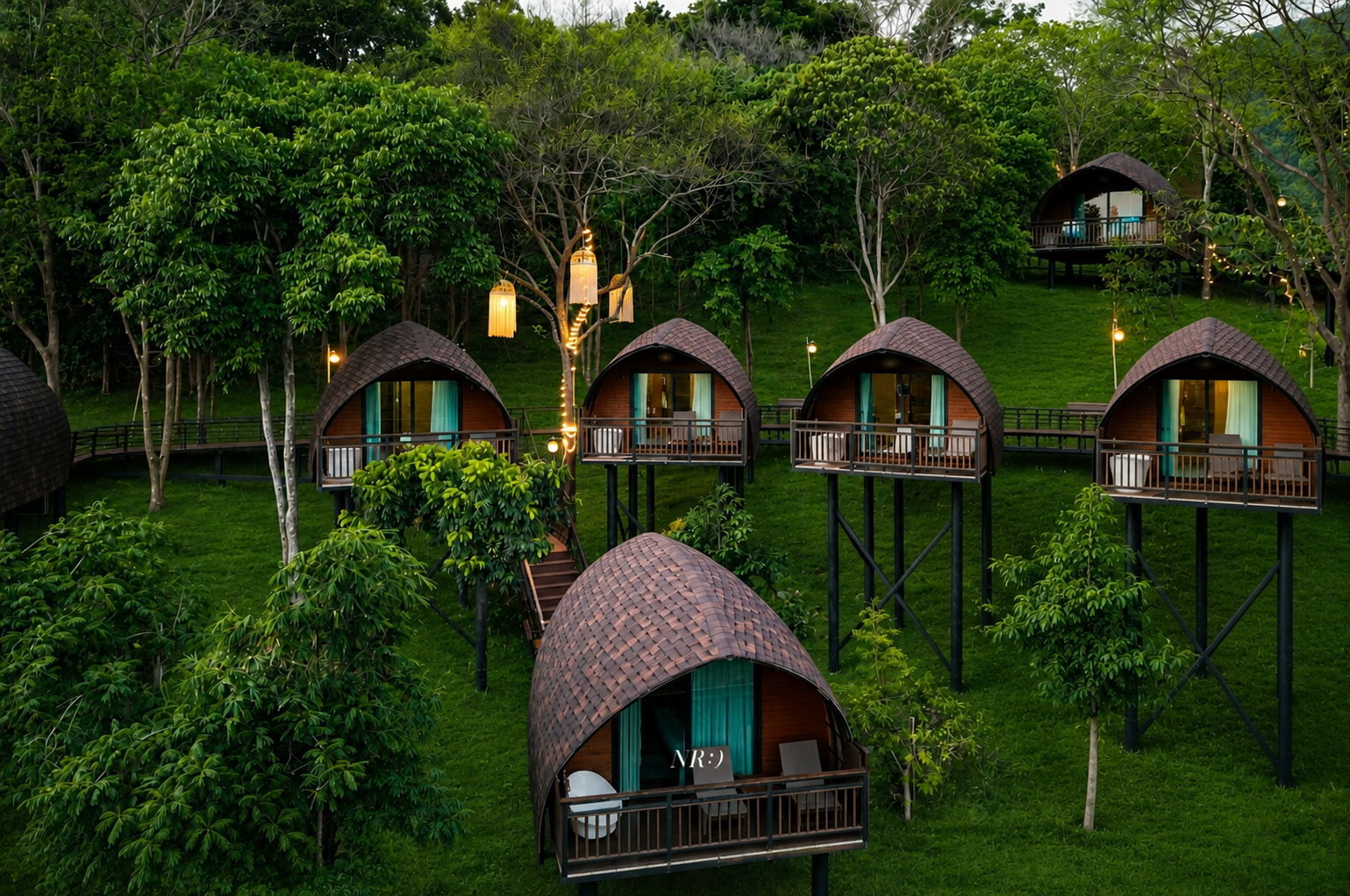Several small, wooden, dome-shaped cabins on stilts in a lush green forest, with string lights hanging from trees, some cabins featuring balconies with outdoor furniture, creating a cozy outdoor retreat setting.