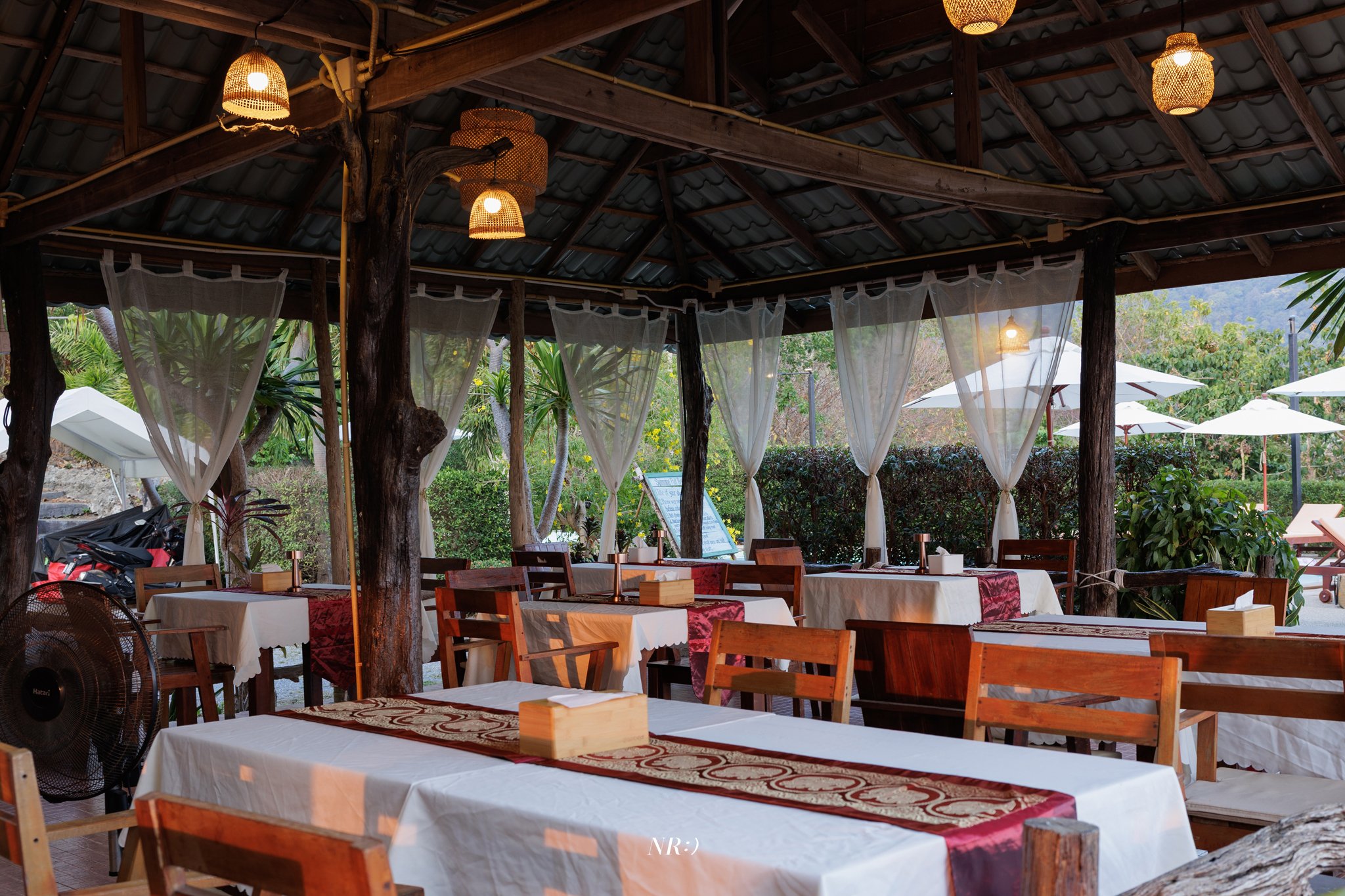 Outdoor restaurant area with wooden tables covered in white tablecloths. Some tables have decorative runners and wooden napkin holders. There are wooden chairs around the tables, sheer white curtains hanging from the wooden beams, and tropical greene