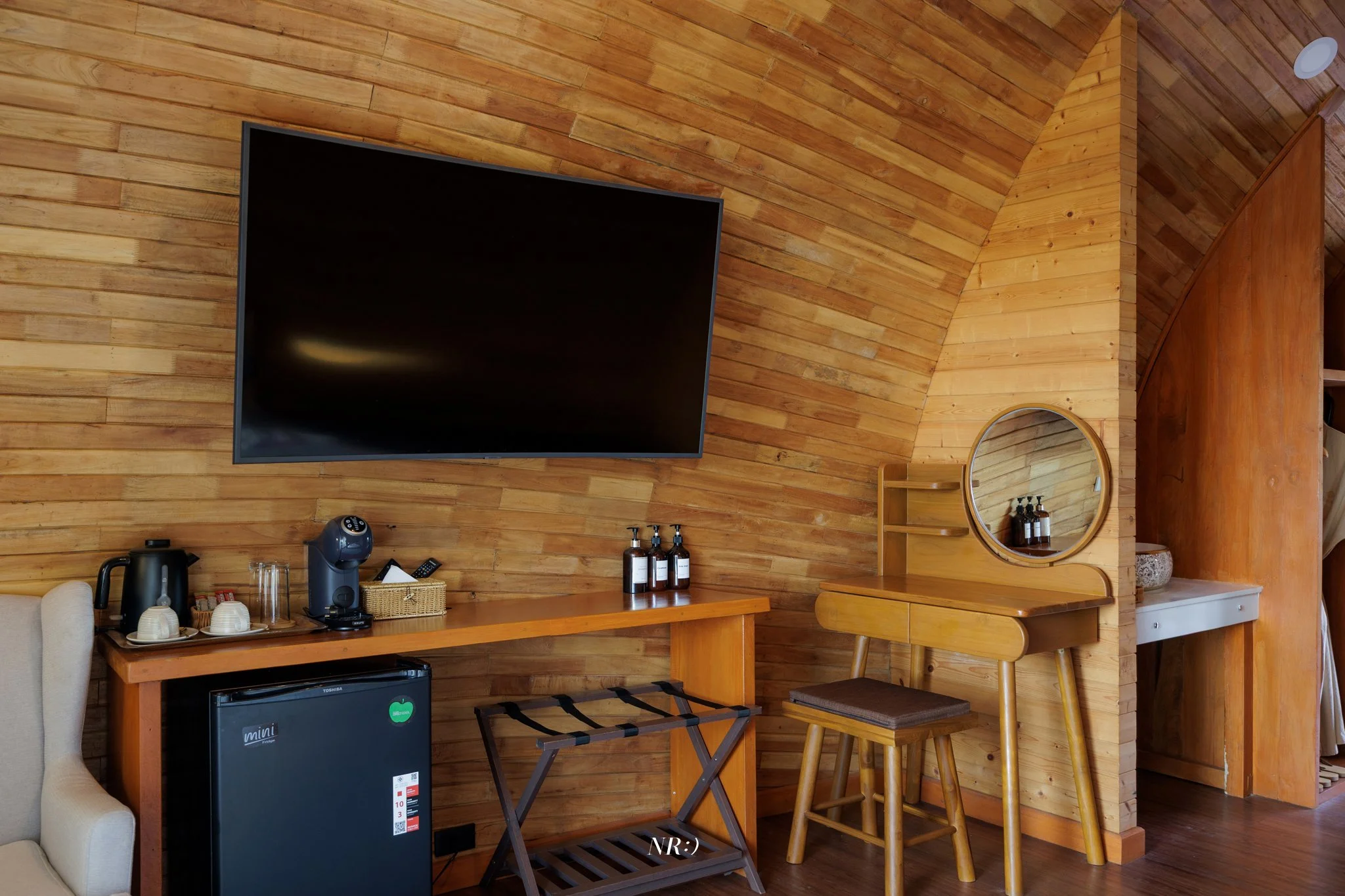 Indoor wooden cabin living area with a wall-mounted flat-screen TV, countertop with coffee maker and cups, small refrigerator, round mirror, and dining table with chairs.