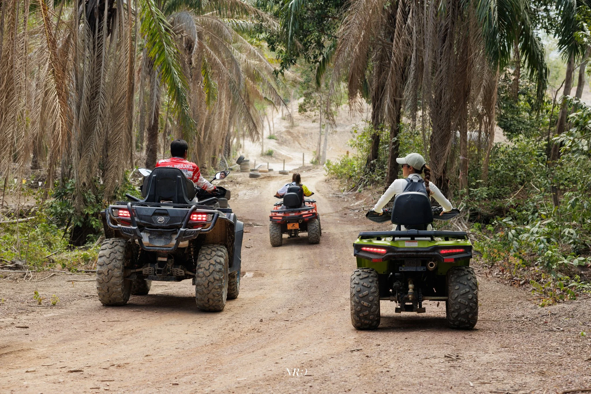 Three people riding all-terrain vehicles down a dirt trail surrounded by tall trees and dense greenery.