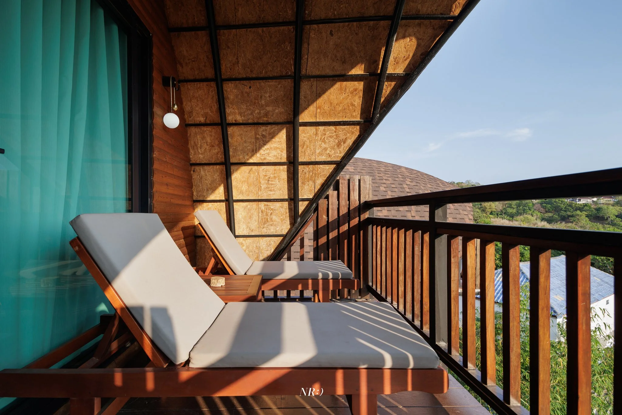 Balcony with two white lounge chairs and a small wooden table. Wooden railing and ceiling, with a scenic view of trees and rooftops in the background. A blue curtain with a round white lamp hanging nearby.