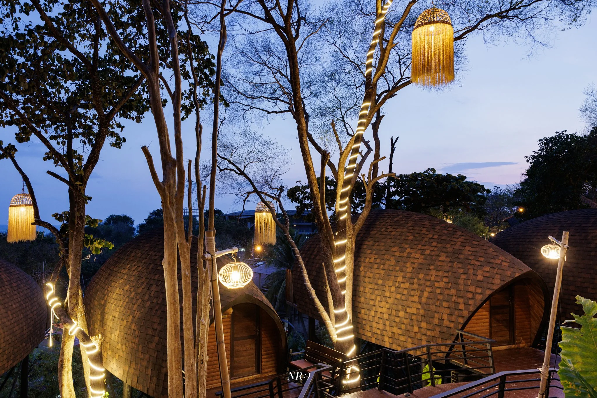 Multiple thatched-roof cabanas with string lights, trees wrapped with string lights, and decorative hanging lanterns, during dusk or early evening.