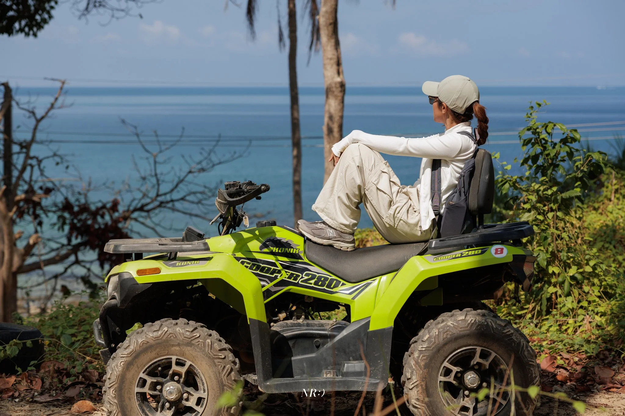 A woman sitting on a yellow ATV near the ocean with palm trees and plants in the background, wearing a beige hat, sunglasses, and casual outdoor clothing.