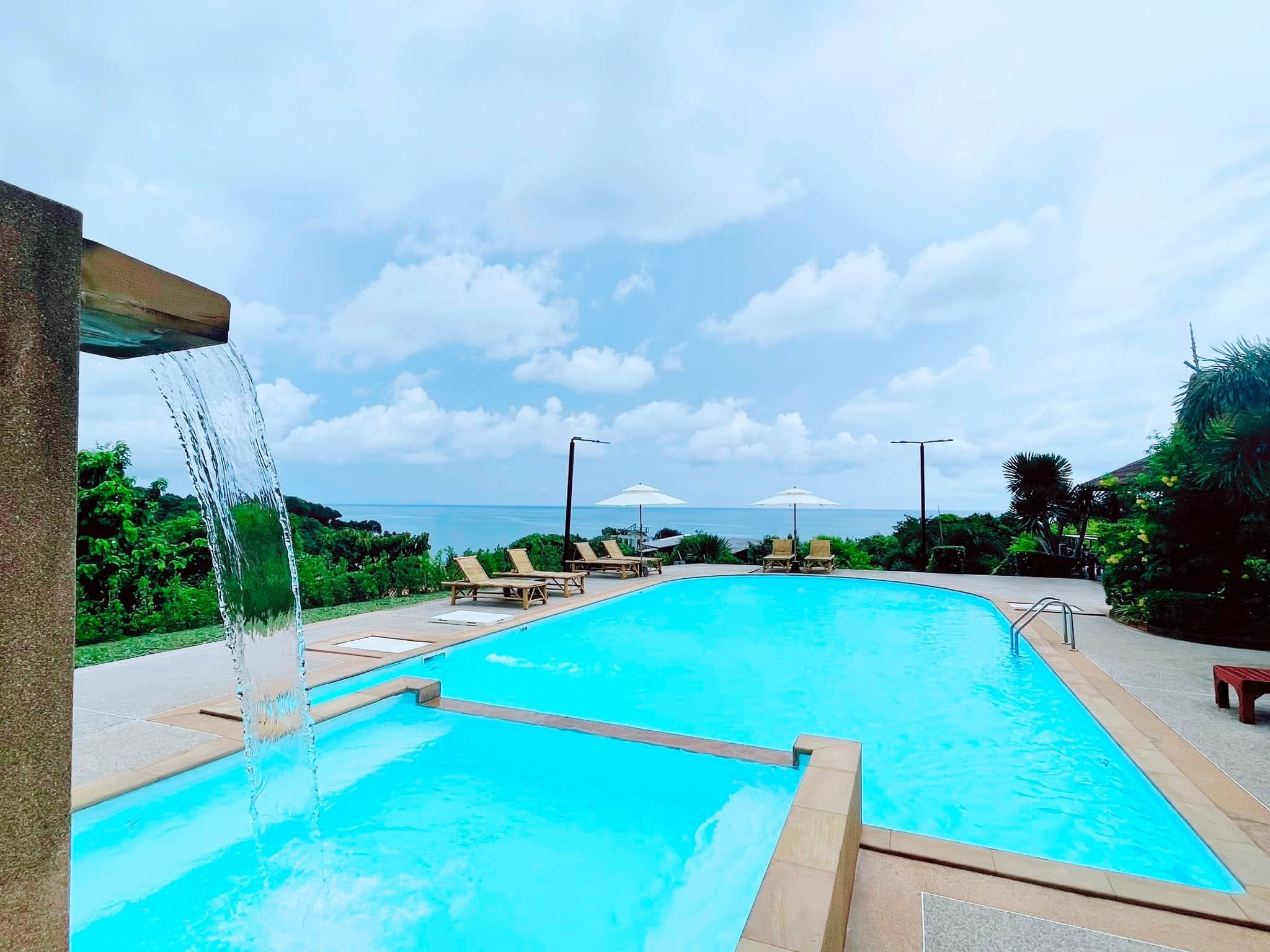 Swimming pool with lounge chairs and umbrellas overlooking the ocean under a partly cloudy sky, with a waterfall feature on the left.