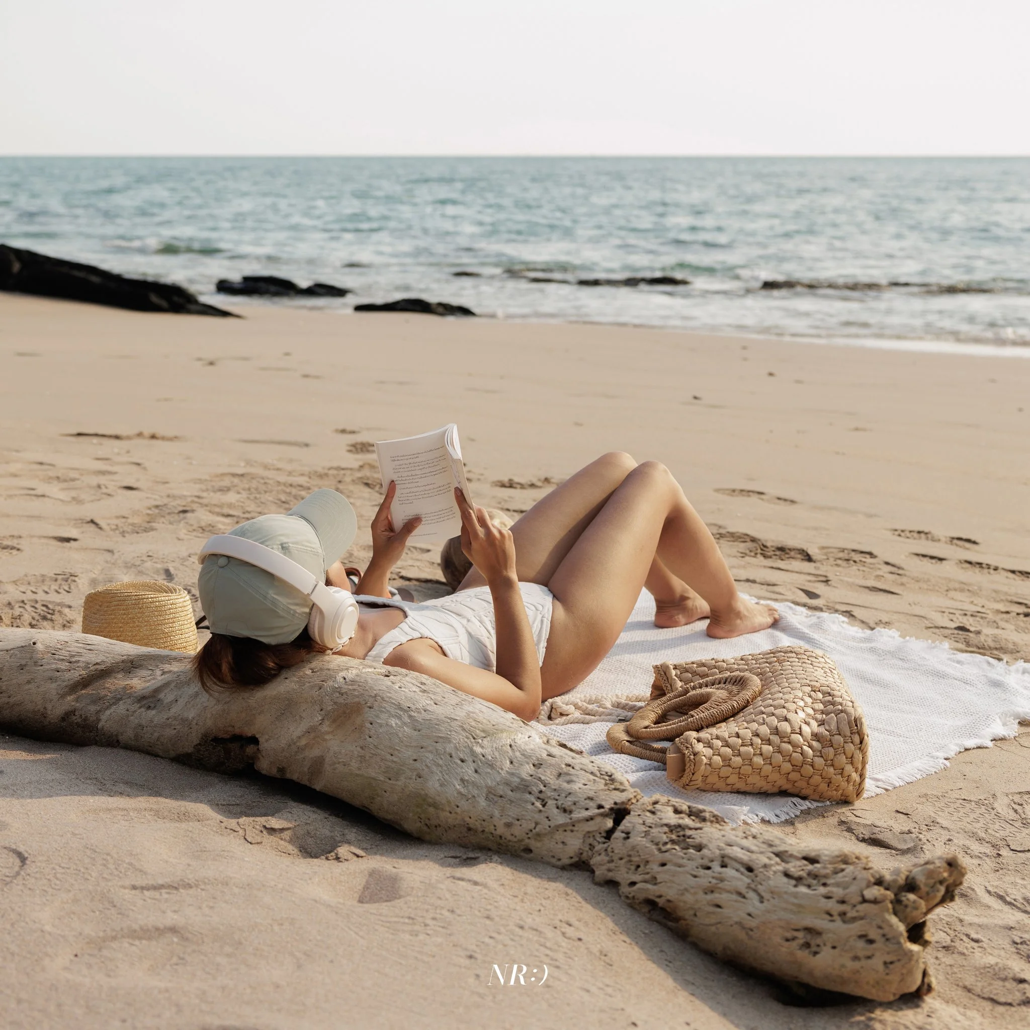  Lying on a sandy beach reading a book with a woven basket and a sun hat nearby, with the ocean and a rocky coastline in the background.