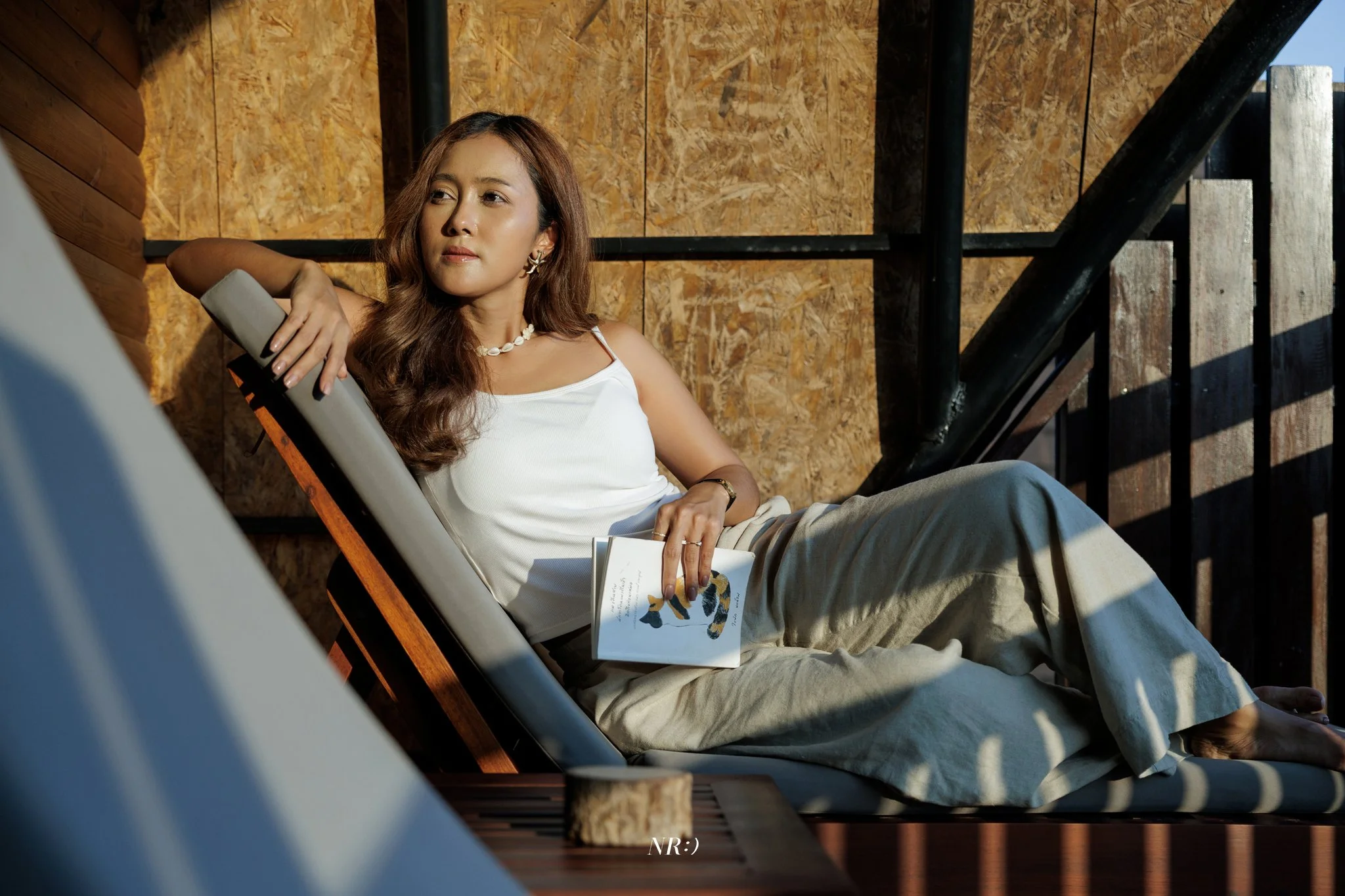 A woman with long brown hair and jewelry sitting outdoors on a wooden lounge chair, holding an open book with dog illustration. She is wearing a white sleeveless top and tan pants, with a wooden background and staircase nearby.
