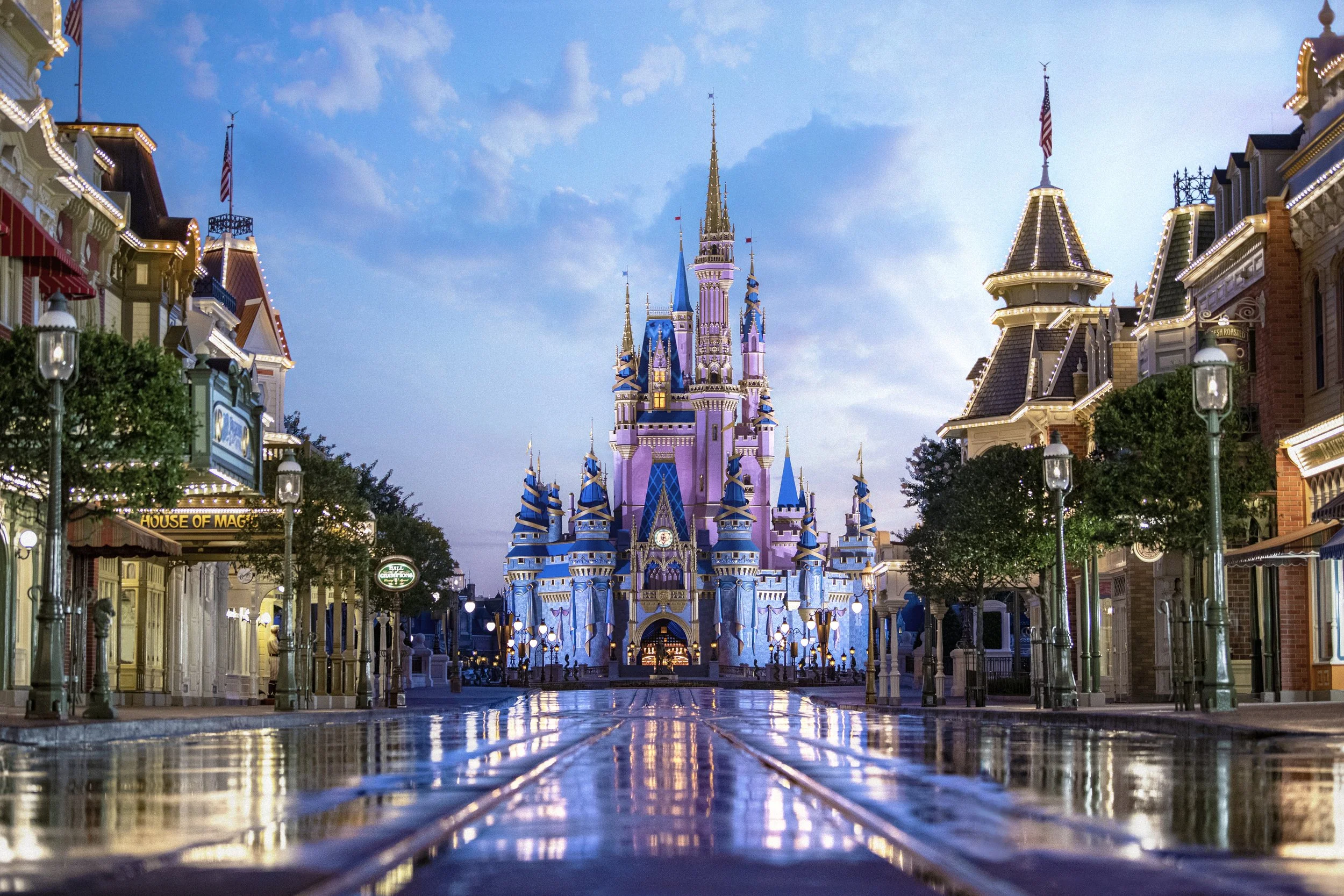 Empty street leading to Sleeping Beauty Castle with wet pavement reflecting lights, surrounded by colorful, historic-style buildings at dusk.