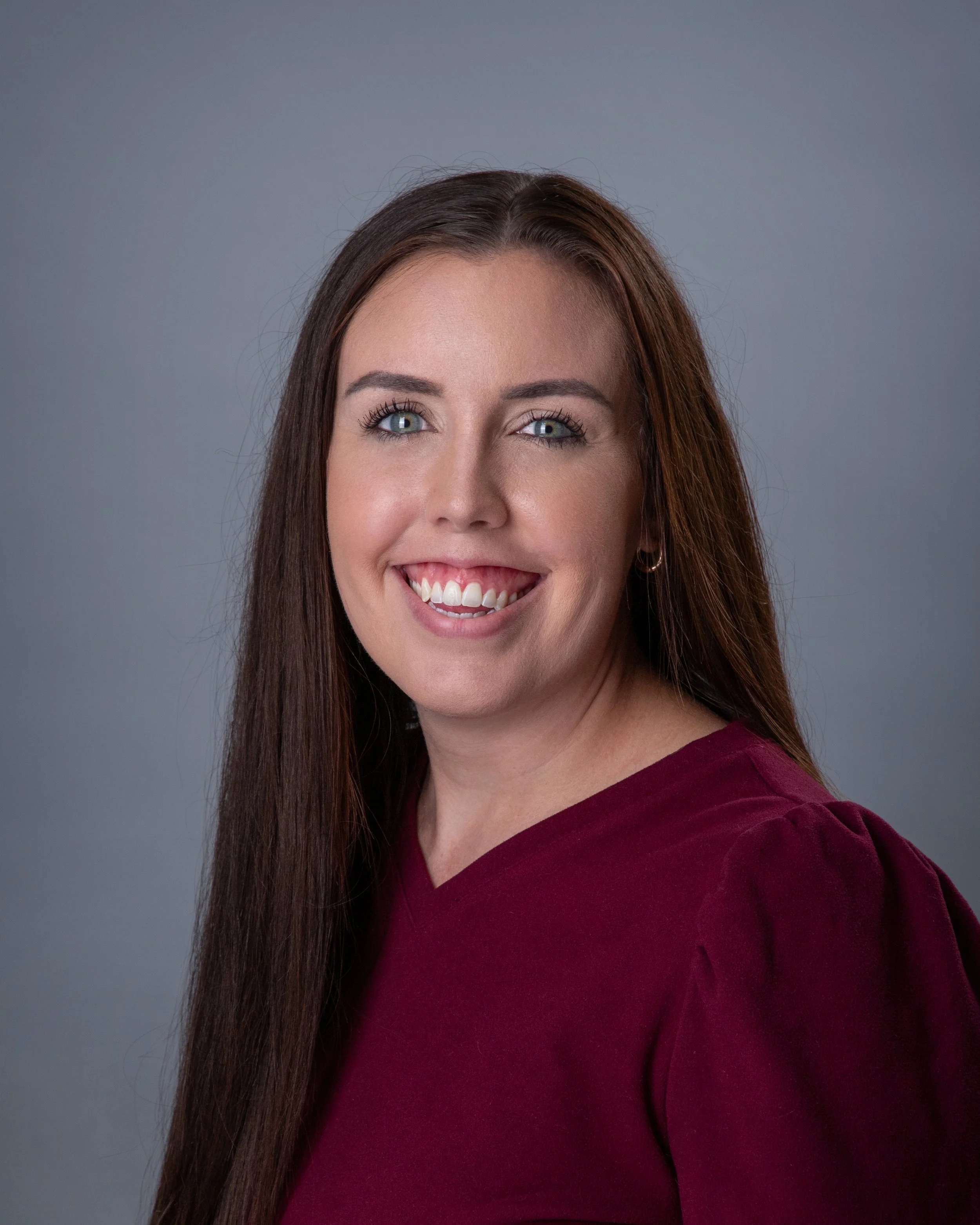A woman with long brown hair, blue eyes, wearing a burgundy top, smiling with teeth, against a plain gray background.