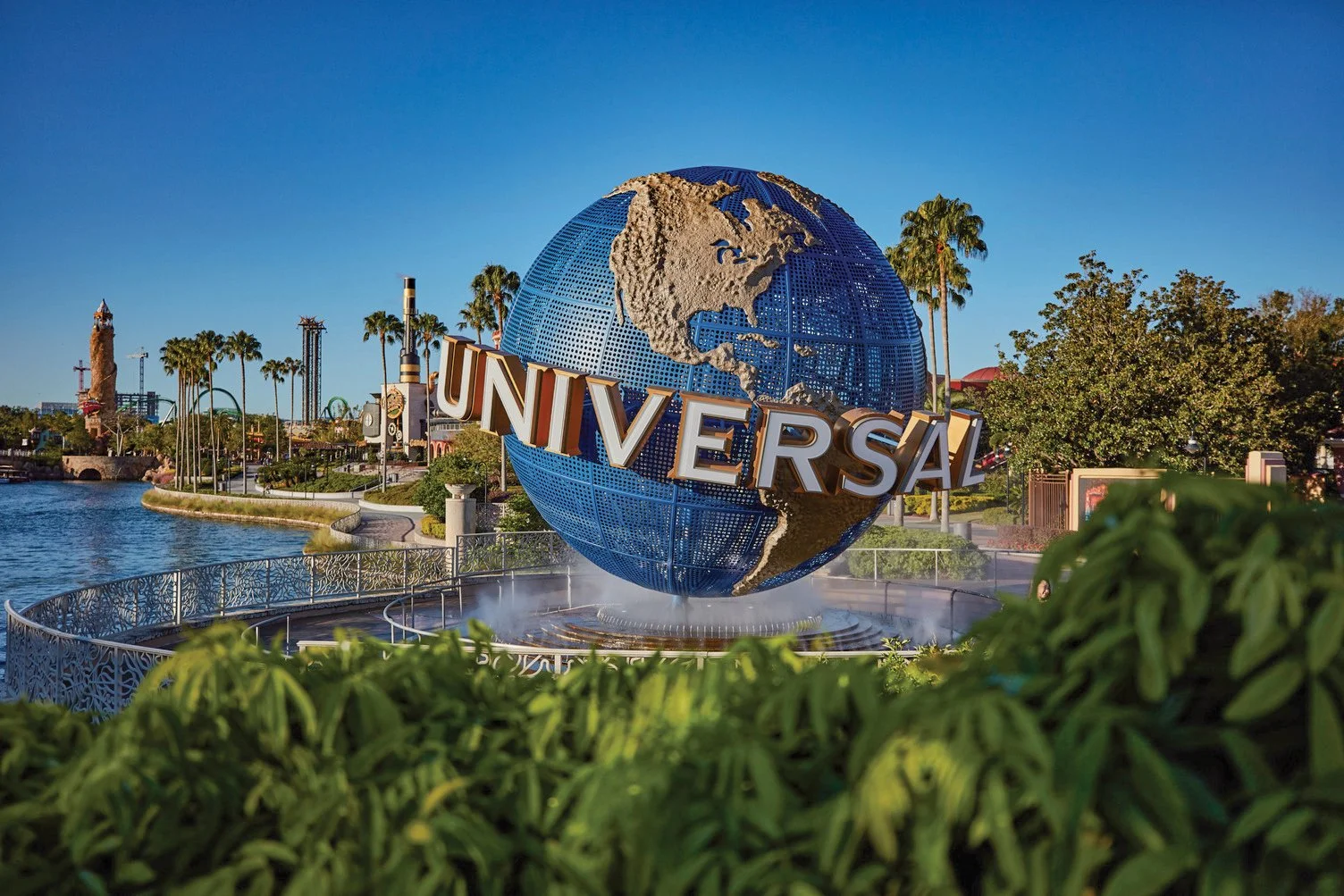Universal Studios globe sculpture at theme park with water fountain in front, surrounded by trees, palm trees, and park rides under a clear blue sky.