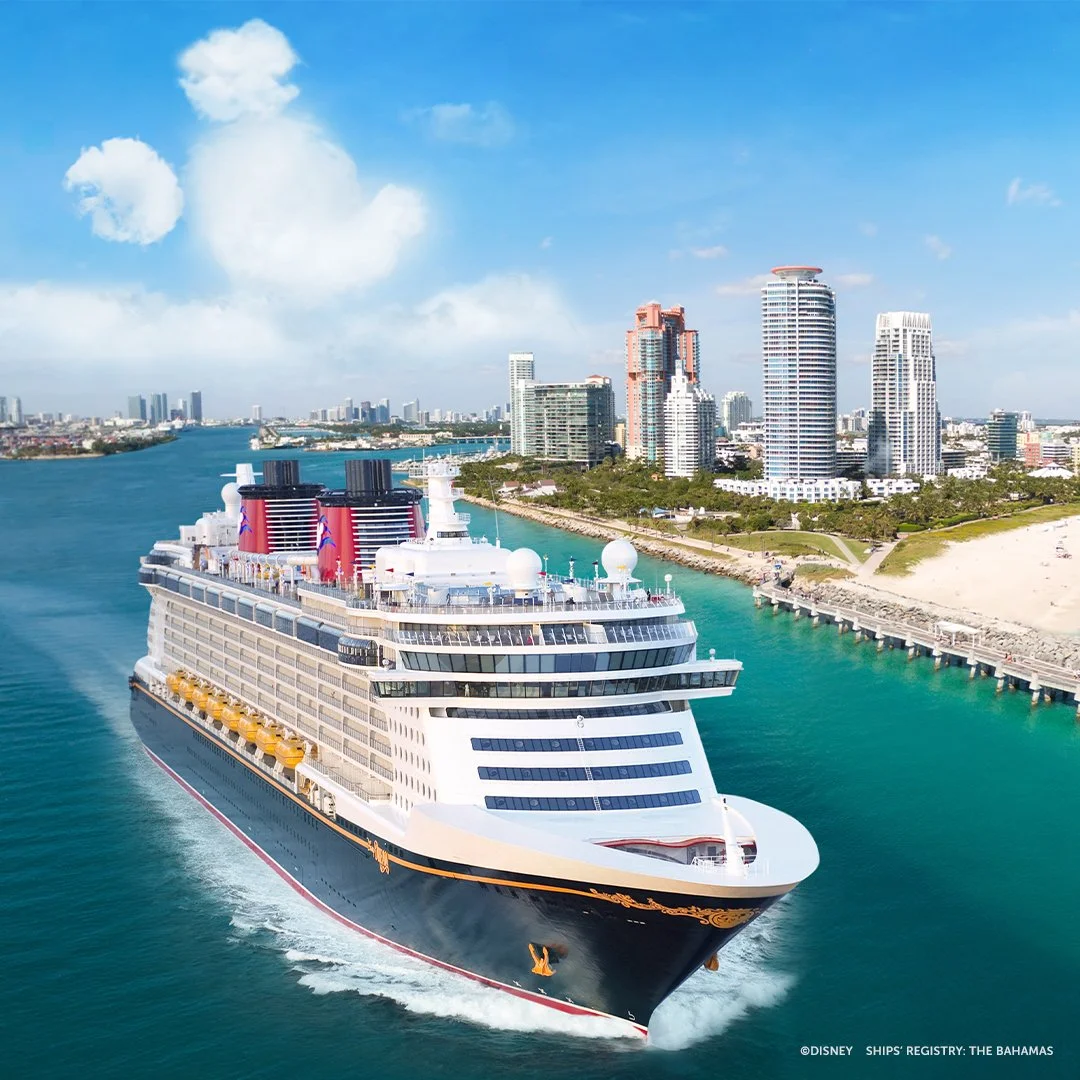 A large cruise ship sailing through a canal near a city with tall modern buildings, blue sky, and some clouds.