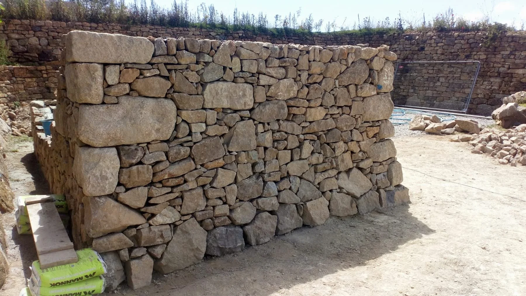 A large dry stone wall made of irregularly shaped rocks stacked without mortar. In the background, there is another similar stone wall and some construction supplies, including bags of mortar and a metal frame.