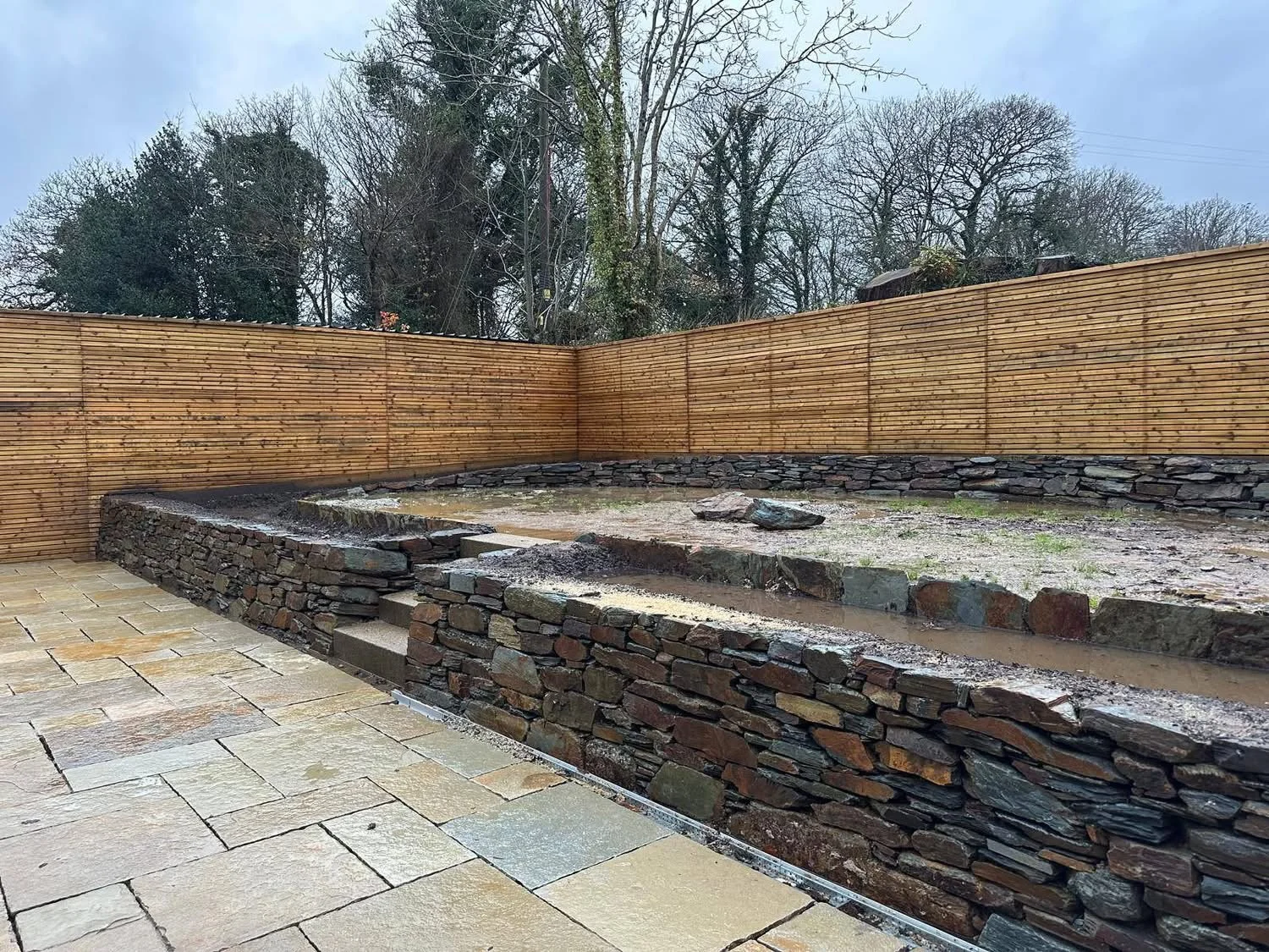 A backyard patio area with an under-construction stone fire pit surrounded by a wooden privacy fence, with trees in the background.