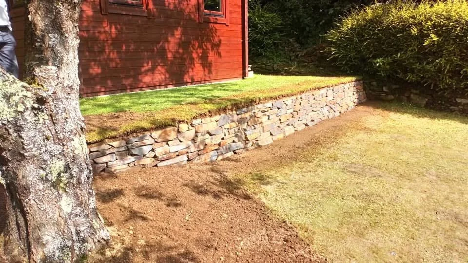 A stone retaining wall supports a raised grassy area beside a red wooden building. A large tree with textured bark is on the left, and a bush is on the right.