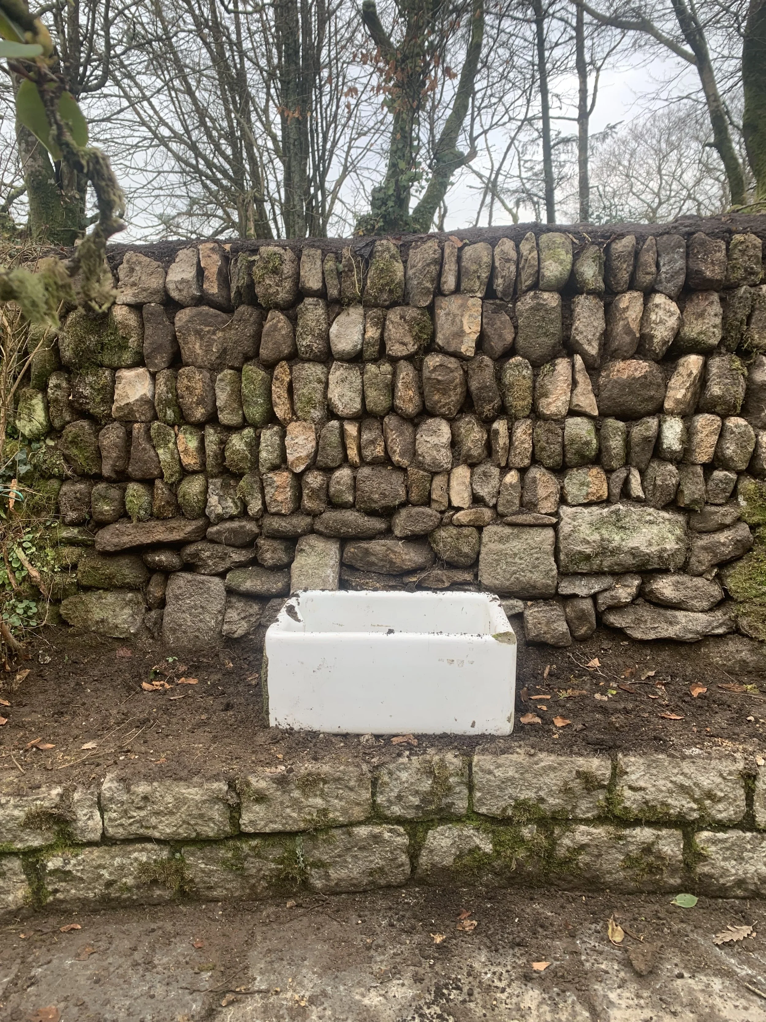 A stone wall with a white rectangular sink attached at the bottom, located outdoors in a wooded area with leafless trees in the background.