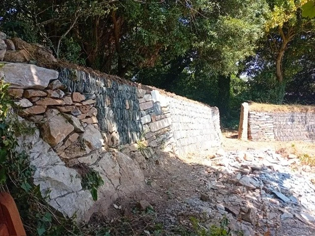 Stone retaining wall alongside a dirt path with trees in background.
