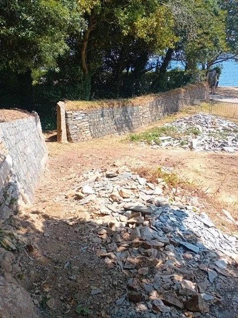 A stone retaining wall with rubble and dirt in the foreground, trees and a person walking on a path in the background.