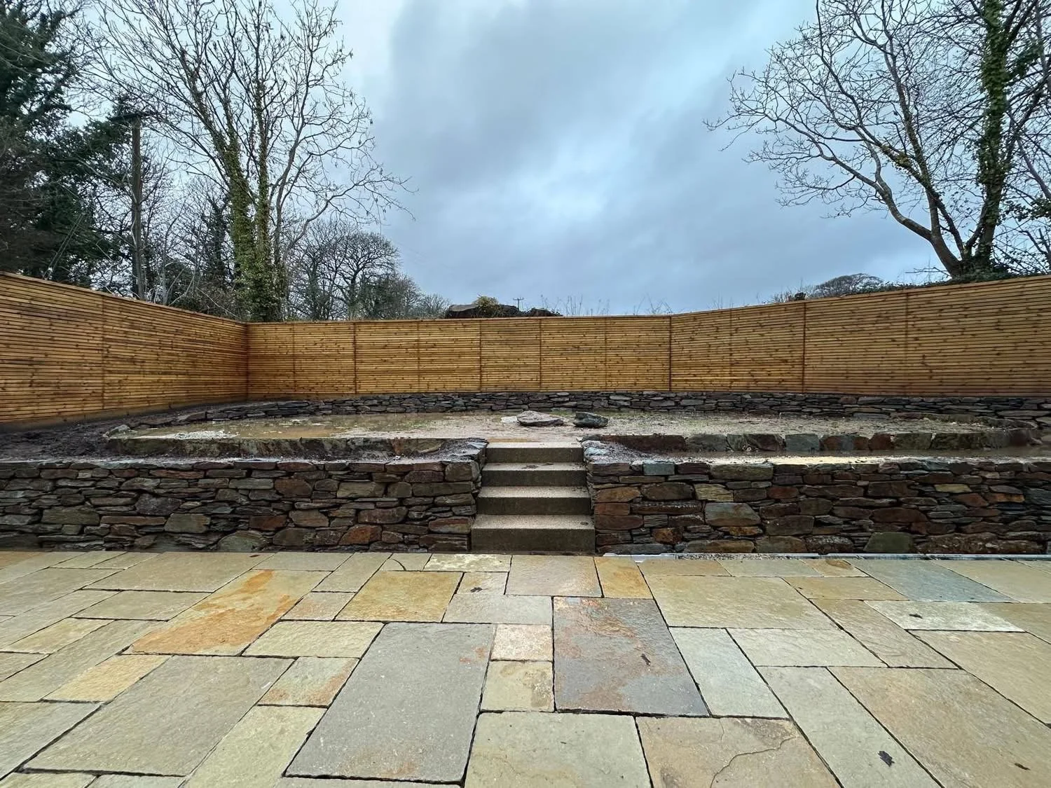 Outdoor backyard with stone patio, steps leading up to a raised area with a stacked stone retaining wall, surrounded by a wooden privacy fence, and several leafless trees against a cloudy sky.