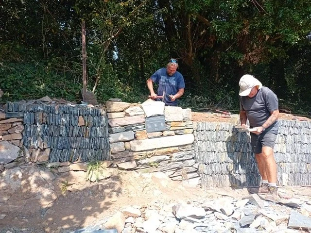 Two men working on building or repairing a stone retaining wall outdoors surrounded by trees, with tools and stones on the ground.