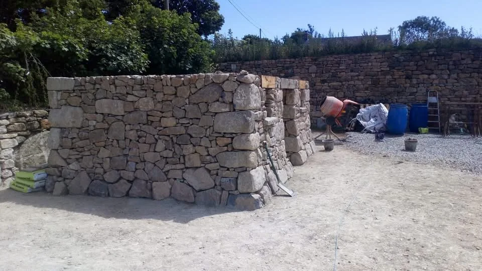A partially built stone wall with construction tools and supplies nearby on a gravel surface under a sunny sky.