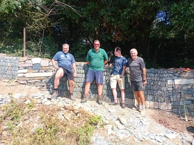 Four men standing on a stone wall outdoors, with trees in the background, posing for a photo.