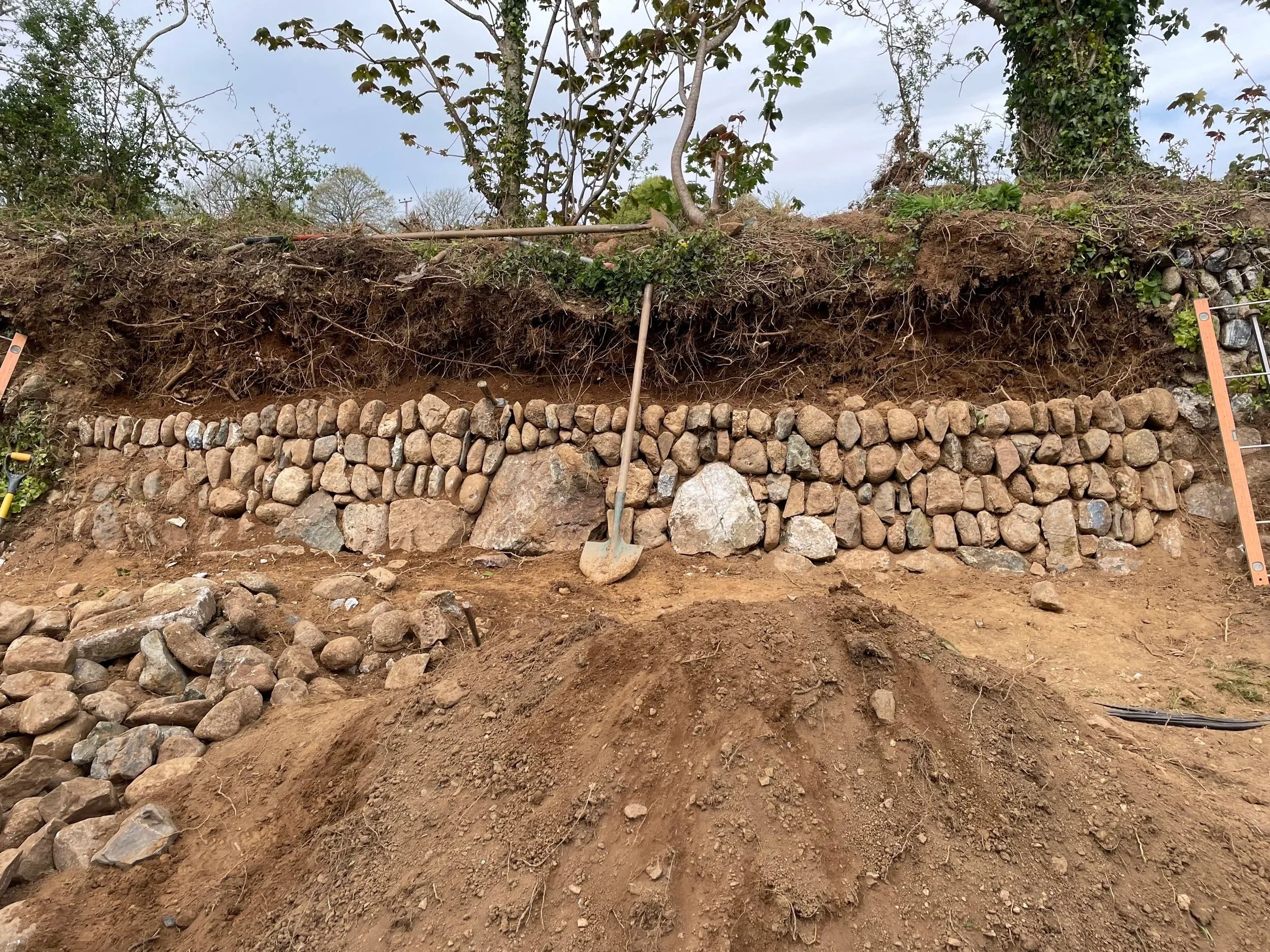 A layered landscape showing a stone retaining wall at the bottom, with soil and roots atop it. Above the soil are plants and small trees, with some gardening tools like a shovel and stake resting against the soil, under an overcast sky.